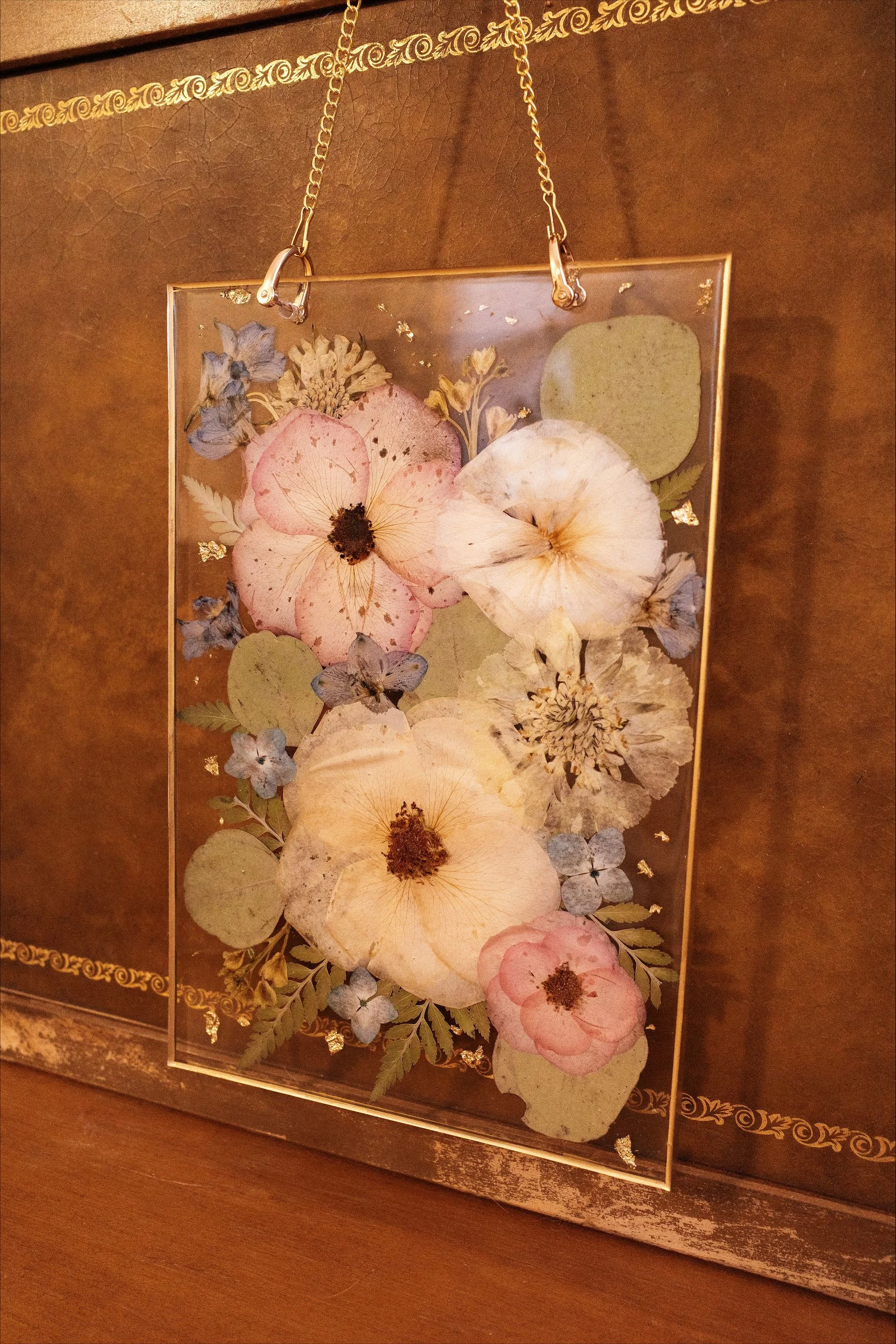 A rectangular glass frame with pressed flowers, attached to a chain hanging from the top, resting against a wooden surface.