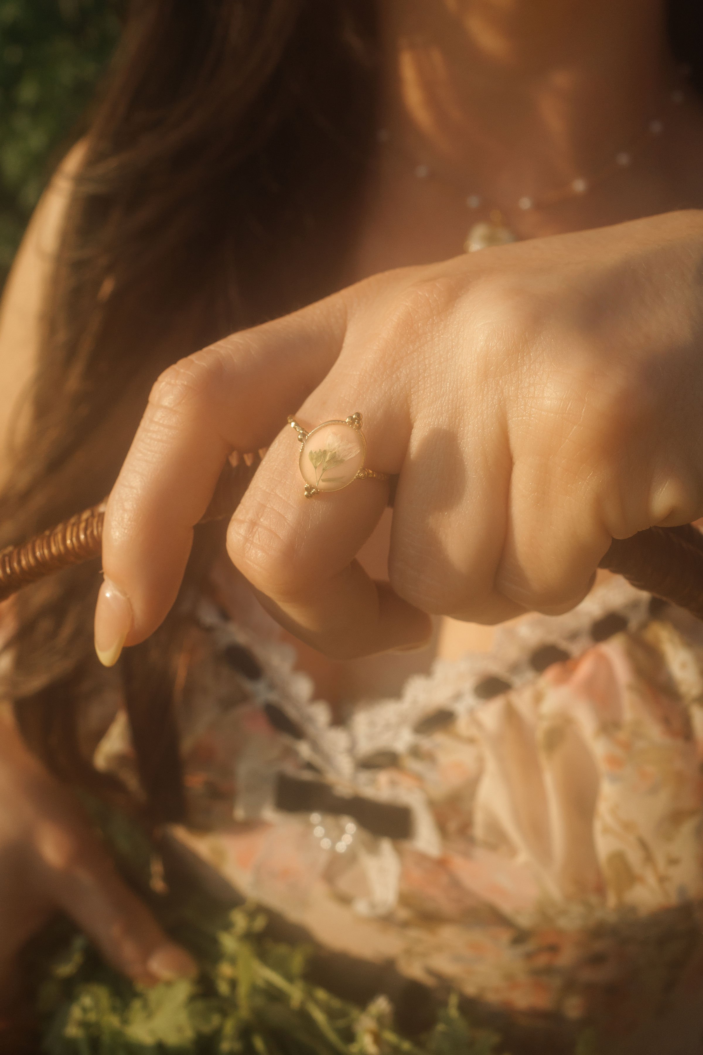 Close-up of a woman's hand wearing a ring with a glass cabochon featuring a flower, with her long, dark hair and a floral patterned dress in the background.
