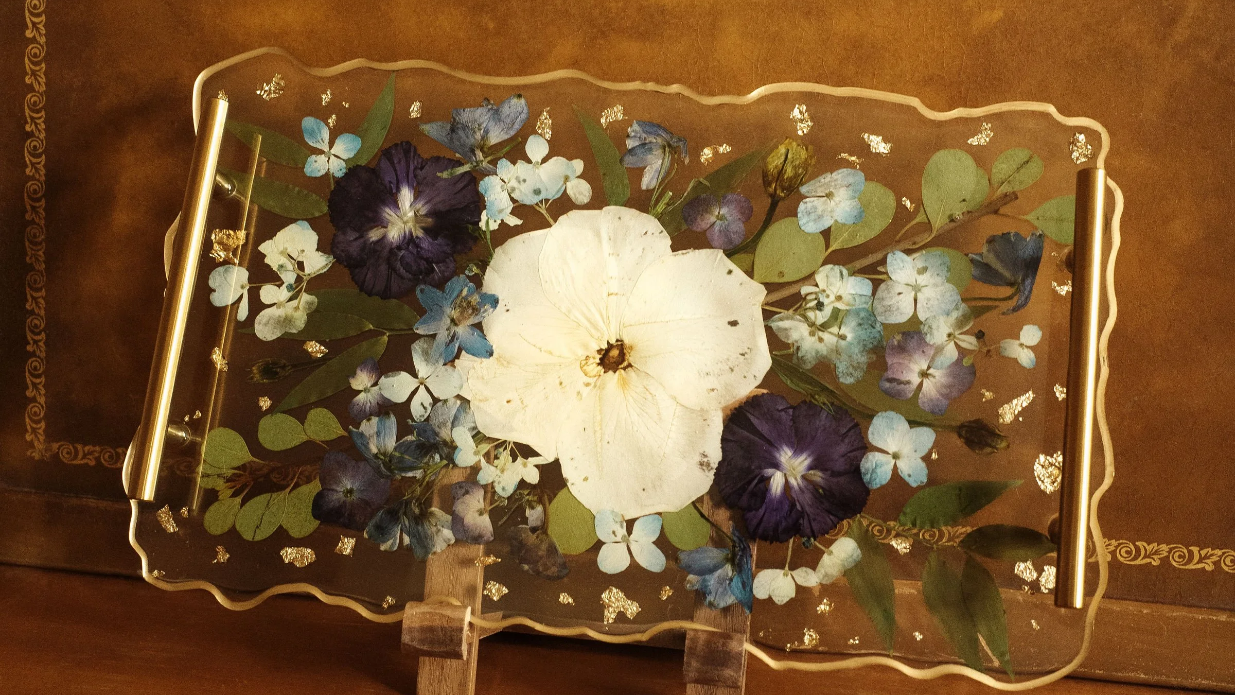 Decorative glass tray with pressed and dried flowers, including white, purple, and blue blossoms, and green leaves. The tray has a wavy gold border and gold handles, displayed on a wooden surface.