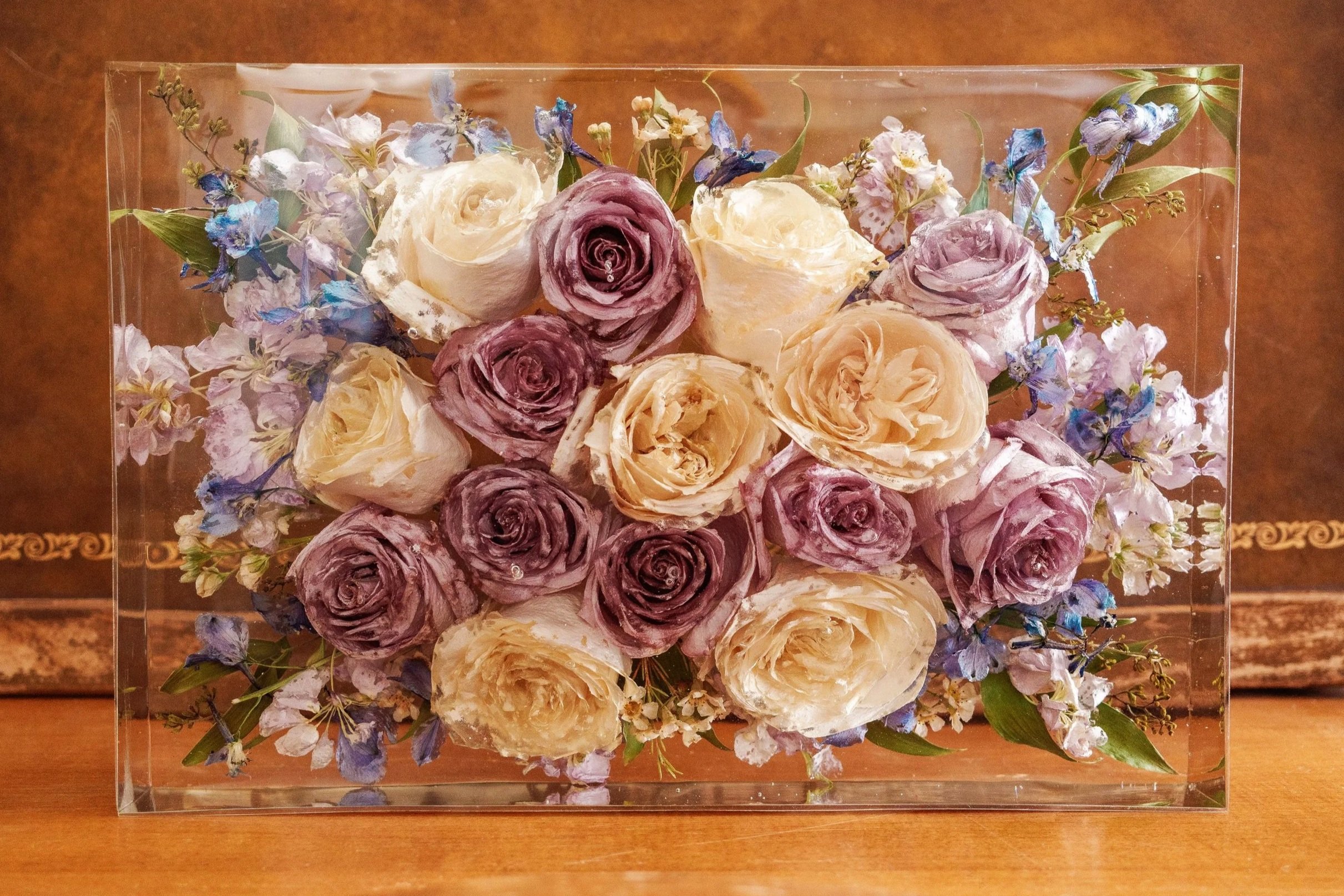 A rectangular glass box containing an arrangement of historical roses in shades of purple, cream, and blush, with small purple and white flowers and green leaves, placed on a wooden surface.