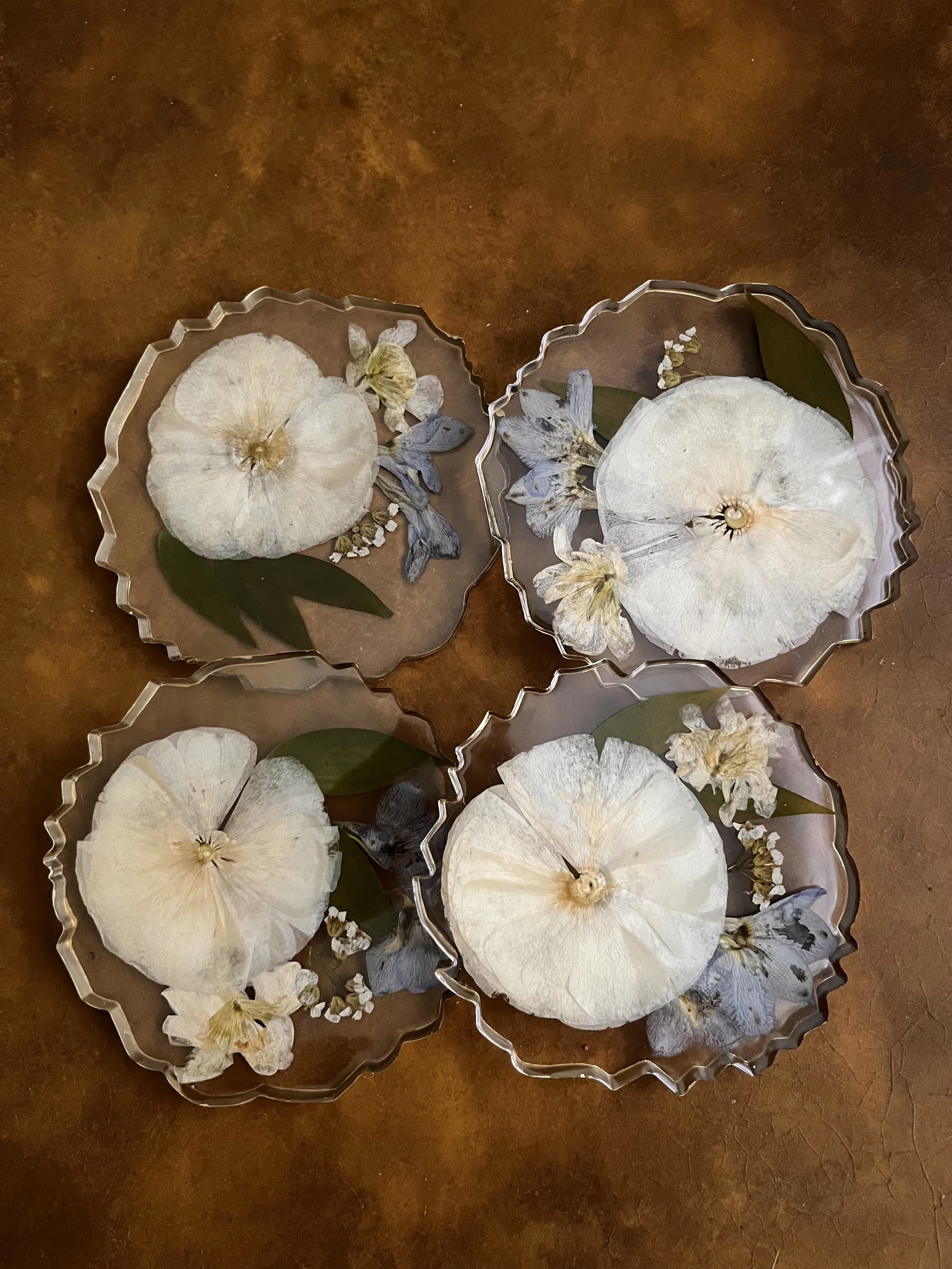 Four glass dishes with dried white flowers, green leaves, and other preserved plant materials on a brown surface.