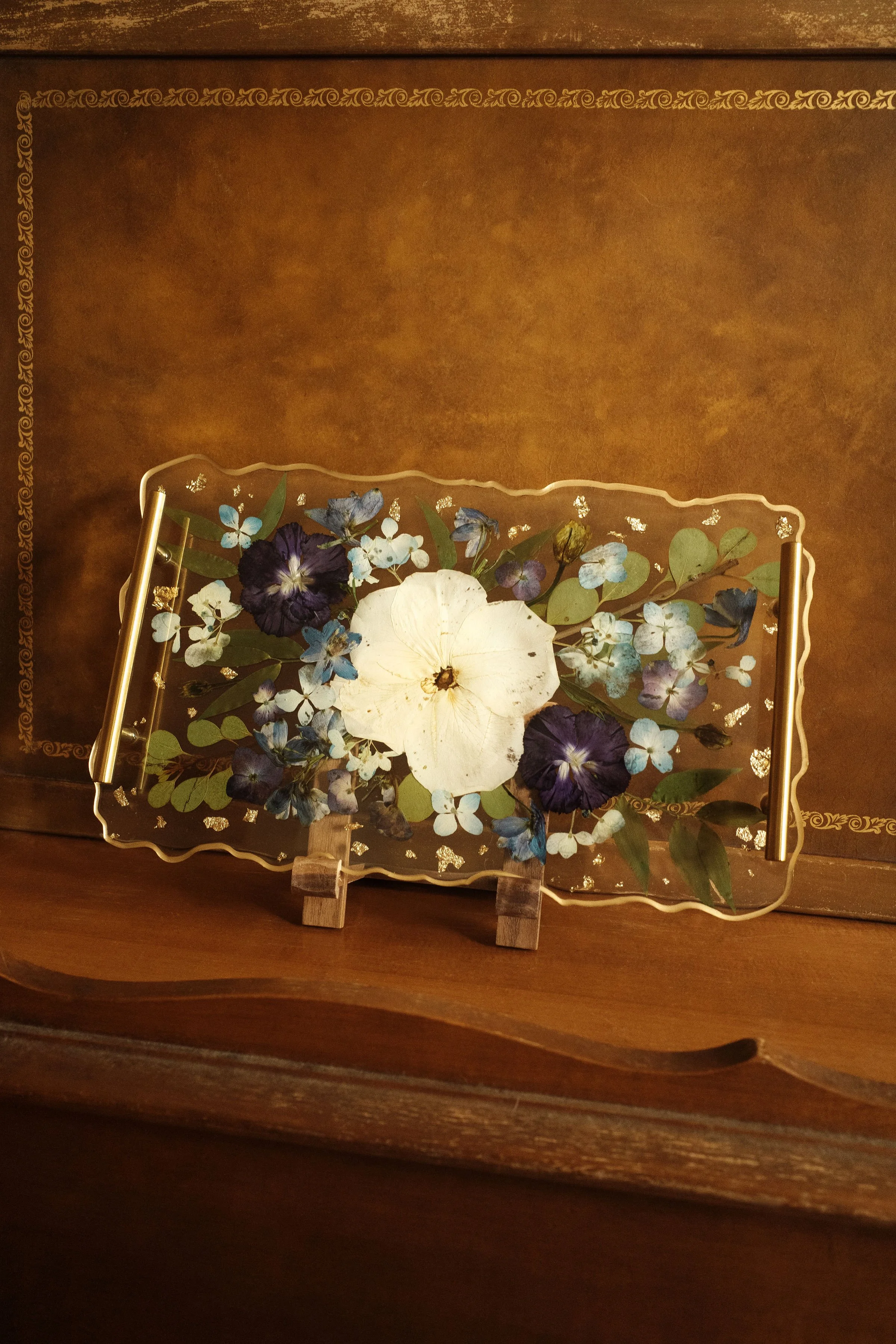 Decorative glass tray with pressed flowers resting on a small wooden stand, placed on a wooden surface against a brown wall with gold decorative borders.