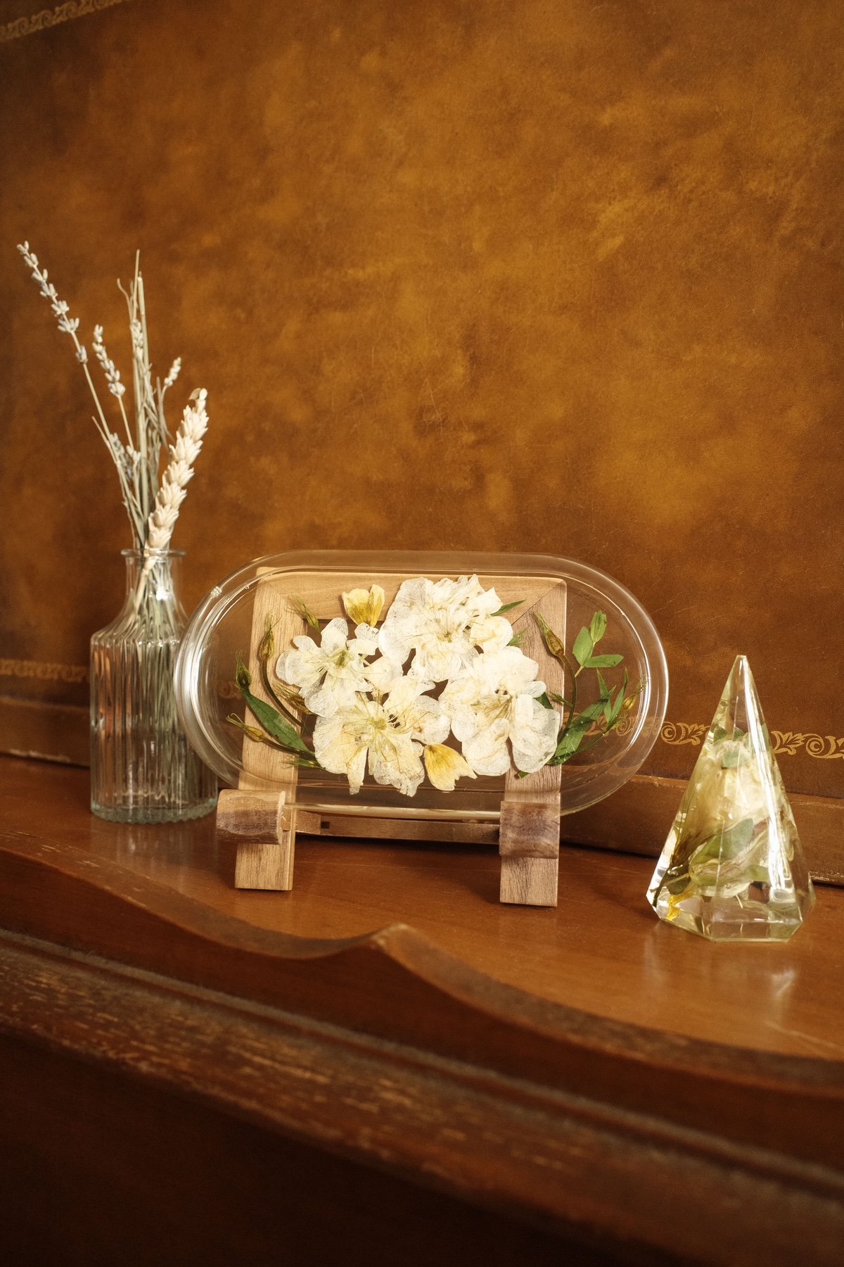 Decorative display with dried flowers in a glass vase, preserved white flowers in a glass container, and a clear glass pyramid, set on a wooden surface against a warm-toned background.