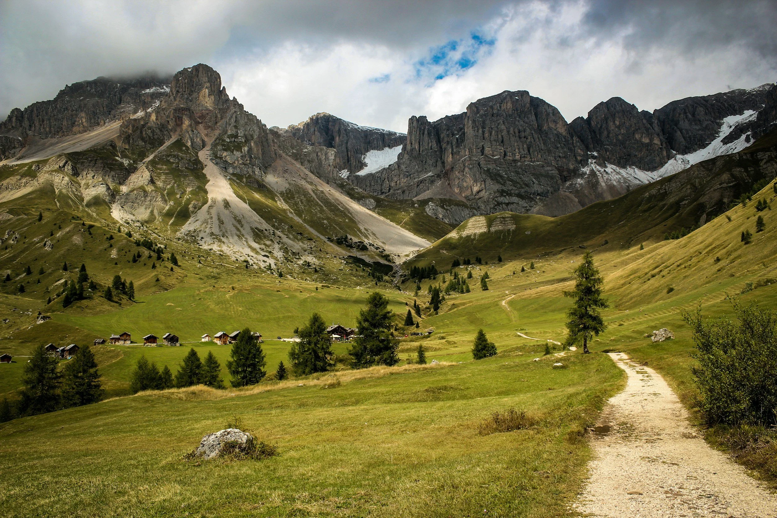 Eine Hügellandschaft mit grünen Wiesen, einigen kleinen Häusern und einem schmalen Weg, vor hohen felsigen Bergen im Hintergrund, teilweise von Wolken bedeckt.