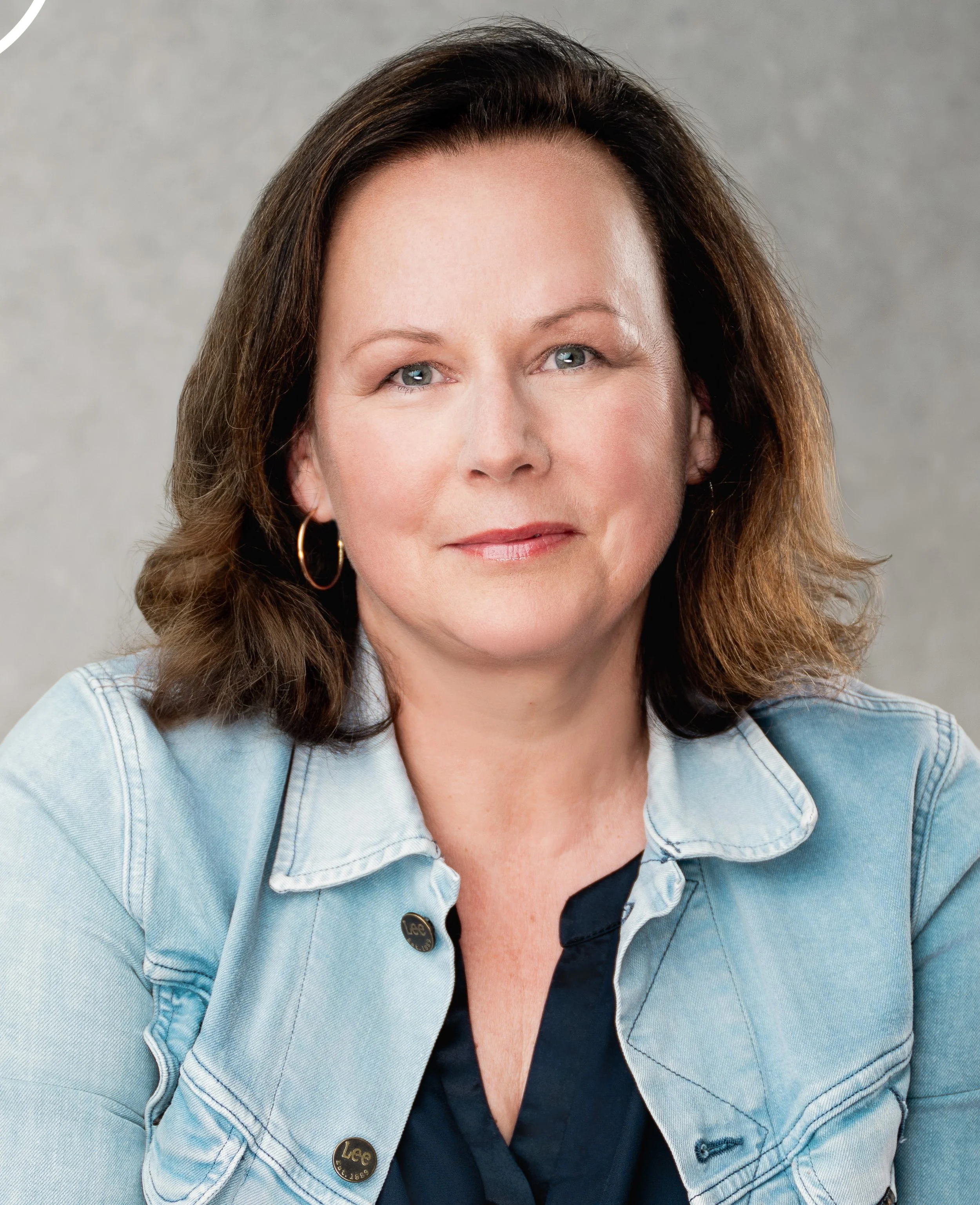 Nahaufnahme der Beraterin Valérie Grass mit braunen Haaren, blauen Augen, in einem dunklen Hemd und einer Jeansjacke, vor grauem Hintergrund.