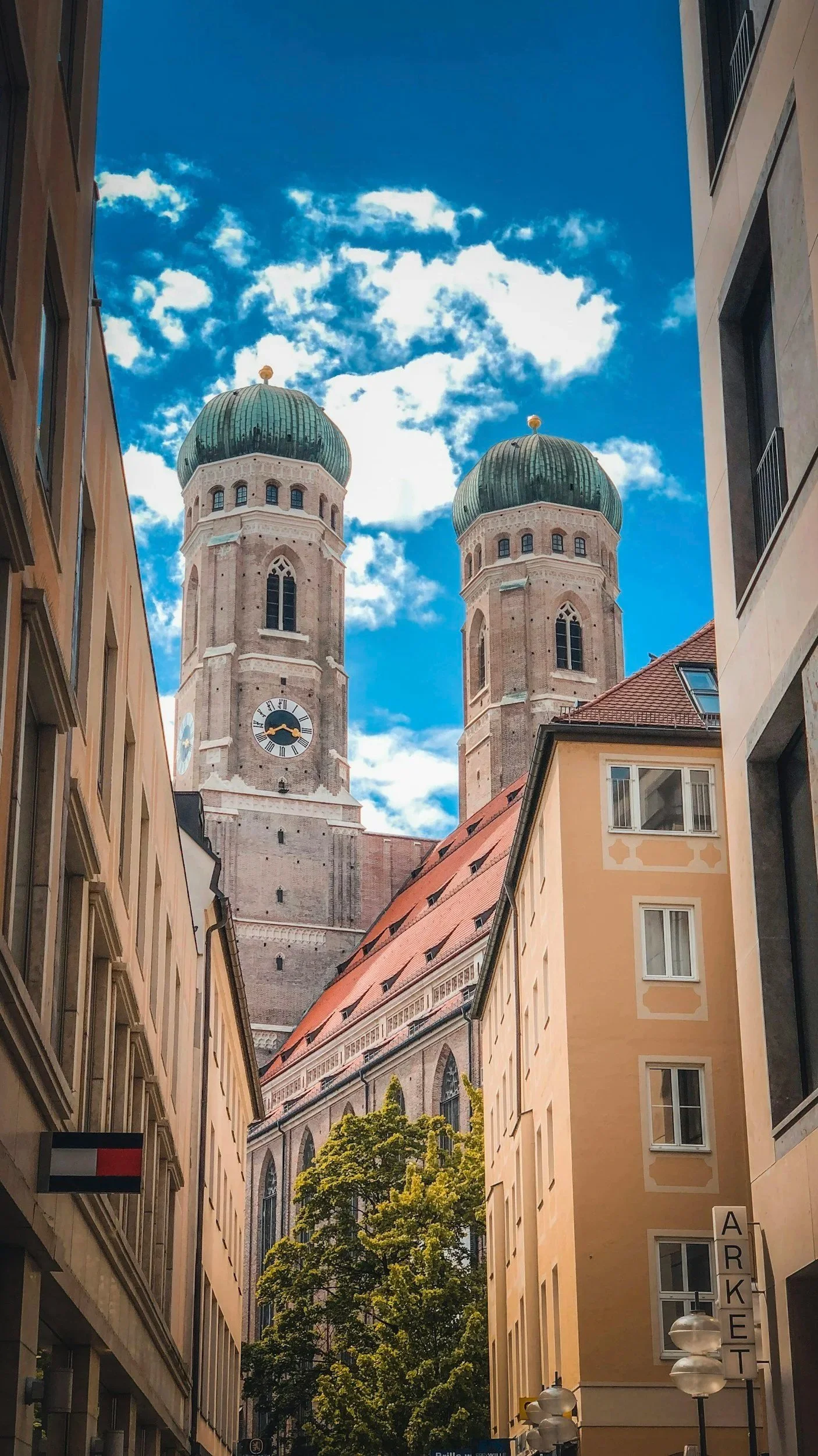 Zwei Kirchentürme mit grünen Kuppeln gegen einen blauen Himmel mit Wolken, umrahmt von Gebäuden in einer Stadt, Blick durch eine schmale Straße.