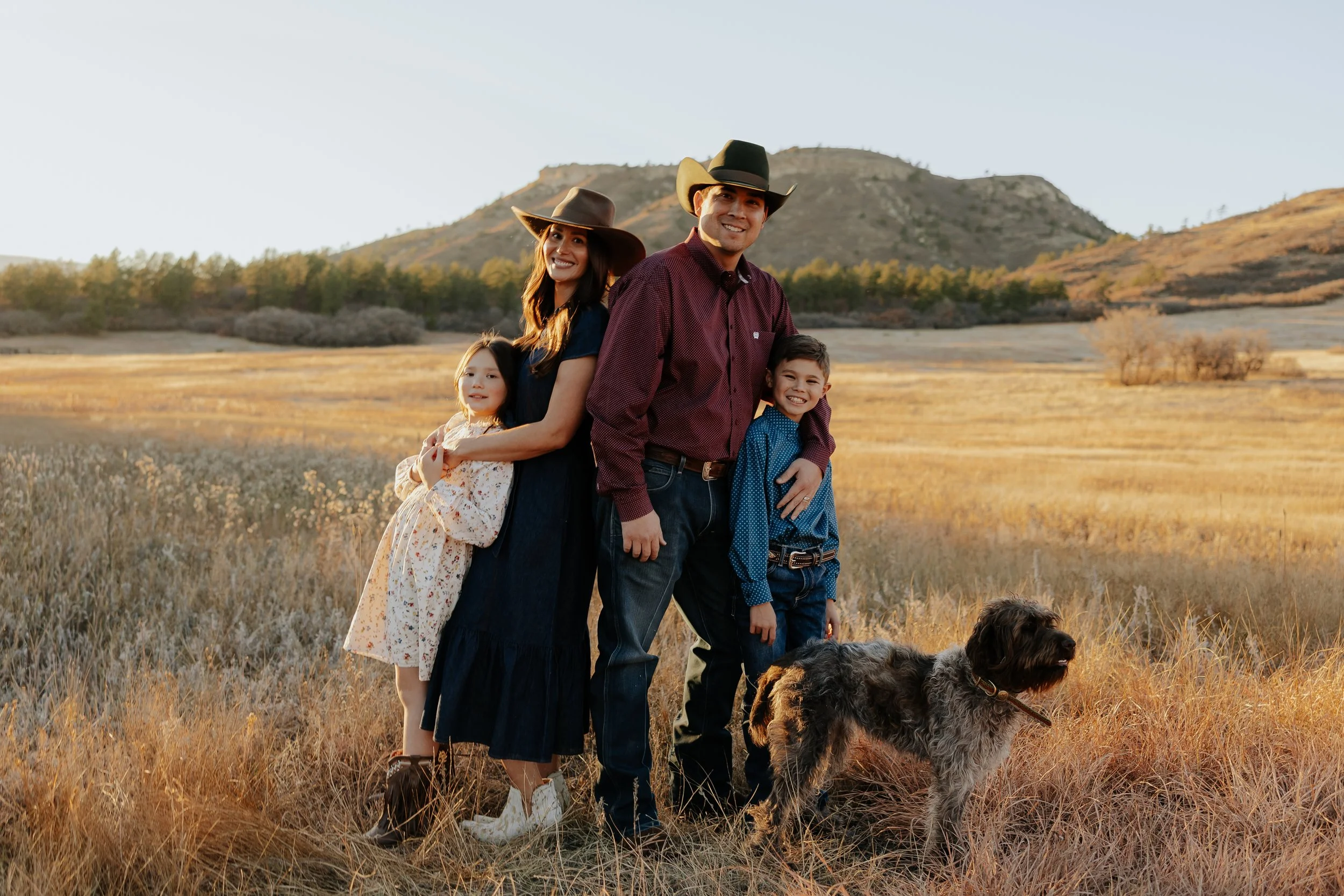 Family of four with a dog standing in a field near mountains during sunset, all smiling, dressed in casual country style with cowboy hats.