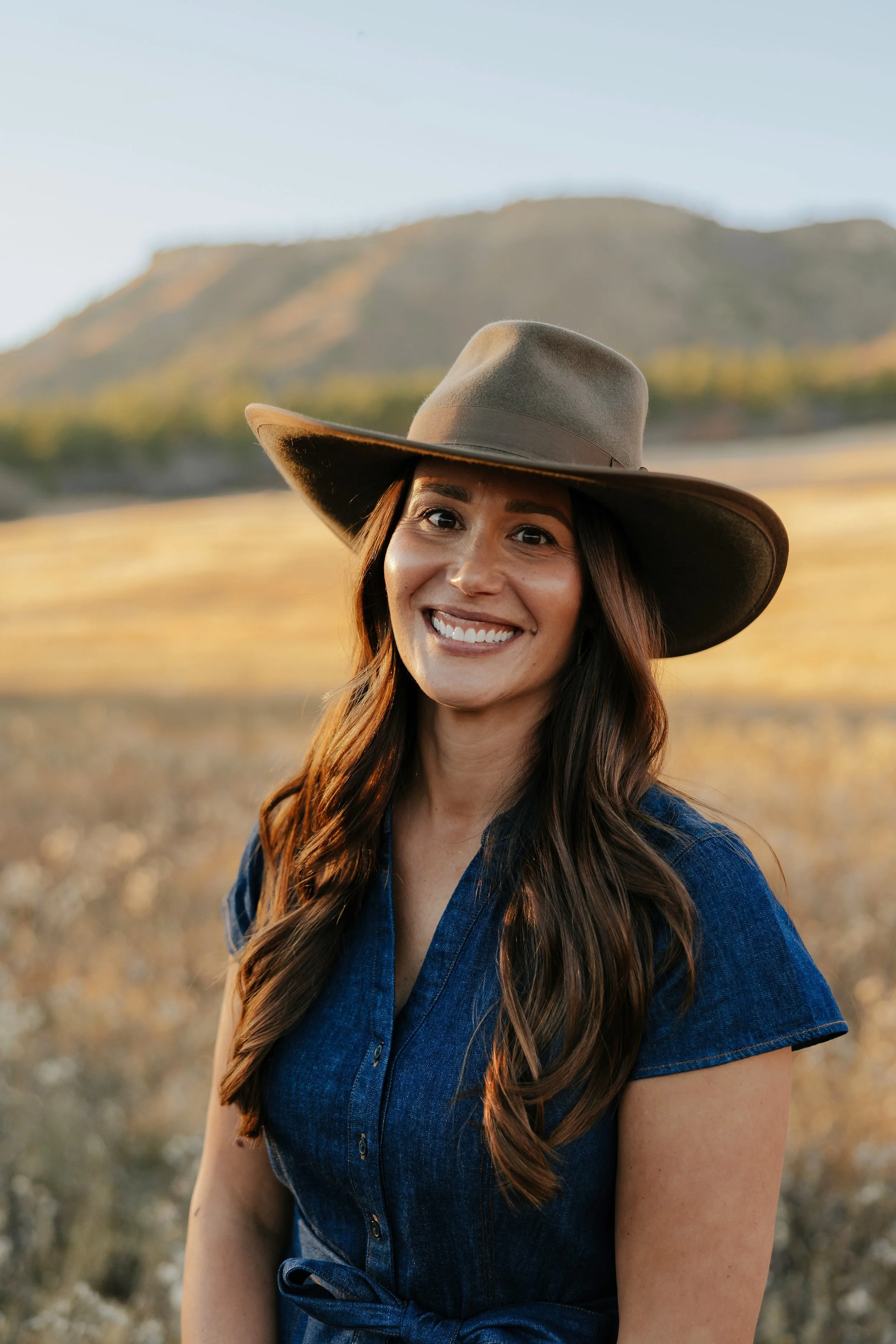 A woman with long brown hair smiling, wearing a wide-brimmed hat and a denim dress, standing outdoors in a field with mountains in the background during sunset.