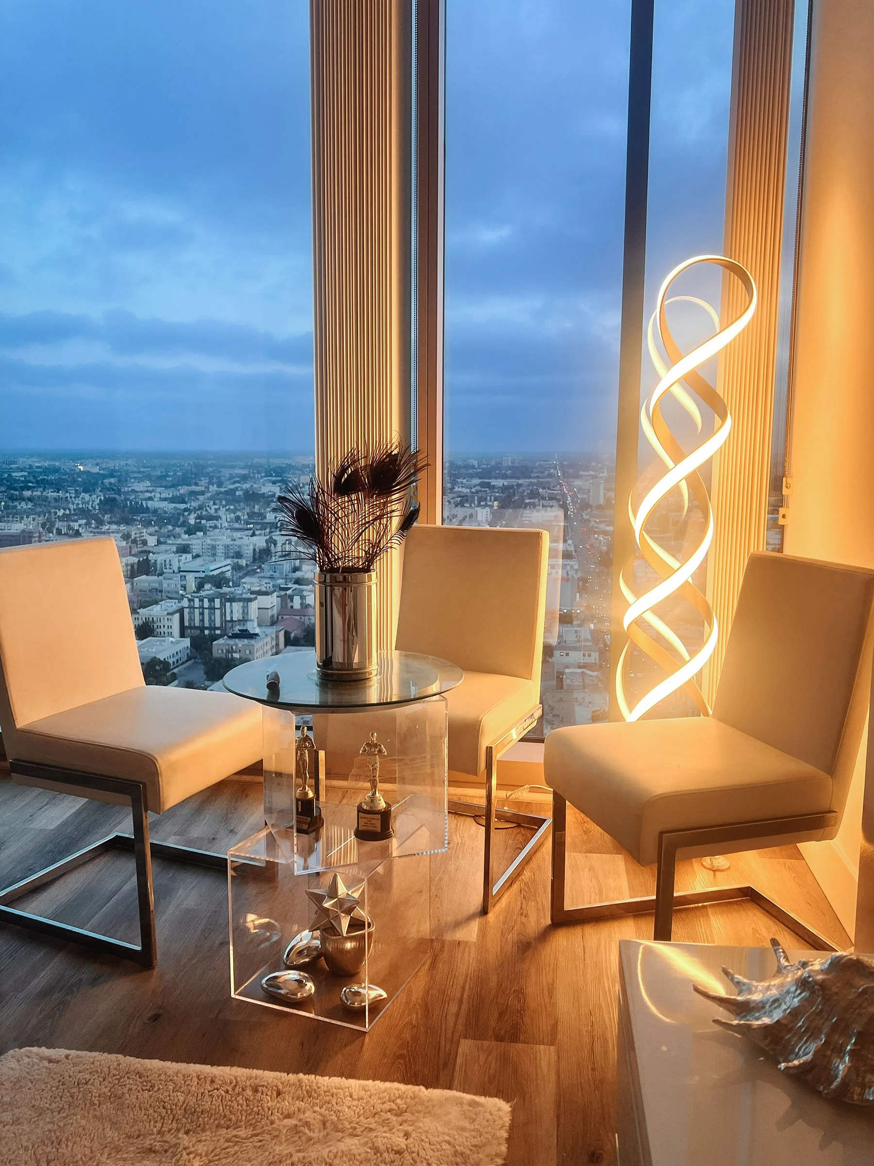 Modern interior of a high-rise apartment or hotel room with floor-to-ceiling windows showing a city skyline at dusk. The room has a glass-topped table, beige chairs, a decorative vase with feathers, and a tall sculptural light fixture.