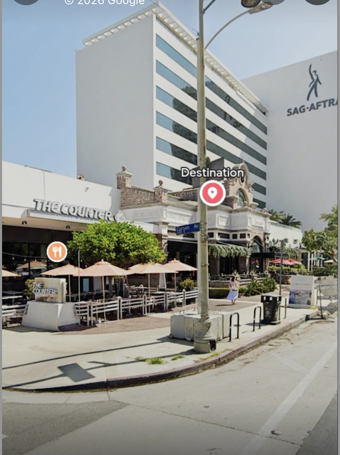 Street corner with outdoor seating area shaded by umbrellas, tall modern building behind with SAG-AFTRA sign, and a historic-style building with decorative architecture, street signs, and a pedestrian in white dress.