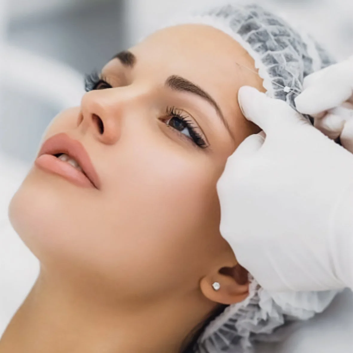 A woman receiving a cosmetic or dermatological treatment on her forehead from a medical professional wearing white gloves.