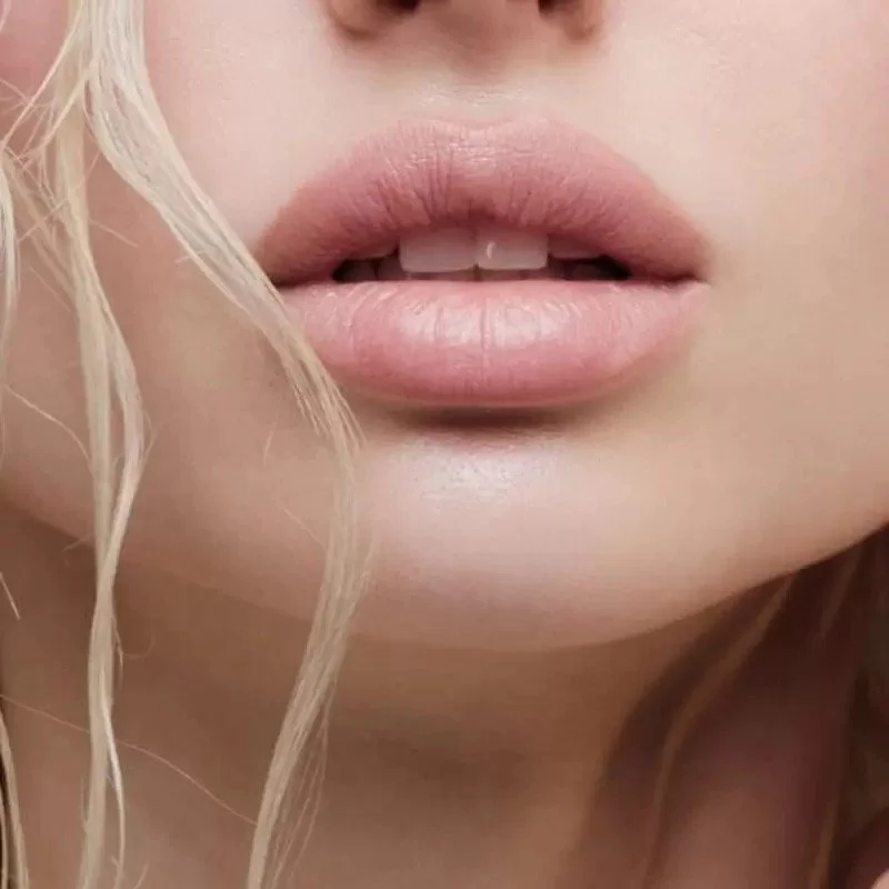 Close-up of a person's lips slightly parted, showing teeth, with light-colored lipstick and blonde, wavy hair.