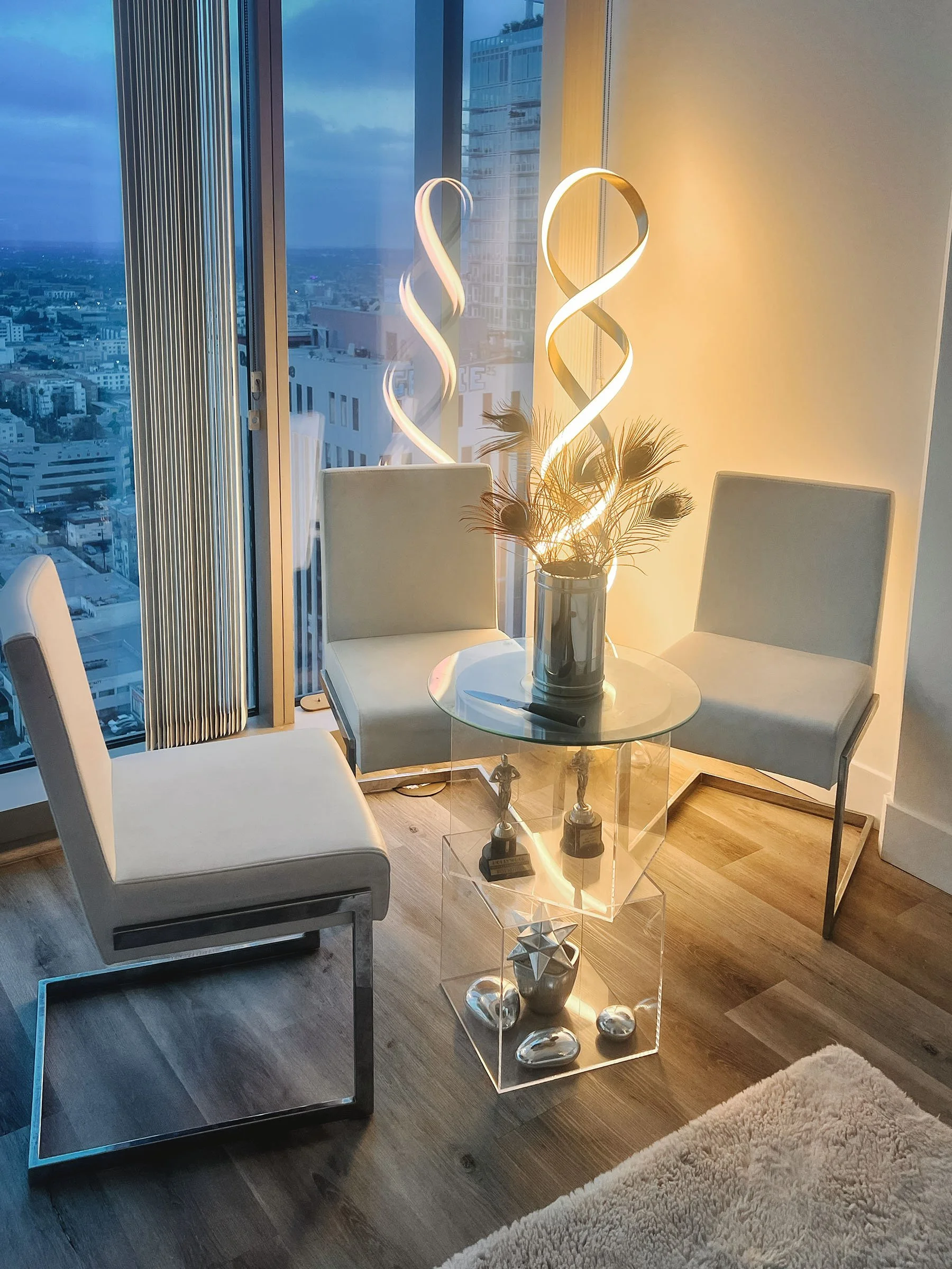 Modern living room with white chairs, a glass side table, decorative objects, an abstract floor lamp, and large windows showing city buildings at dusk.