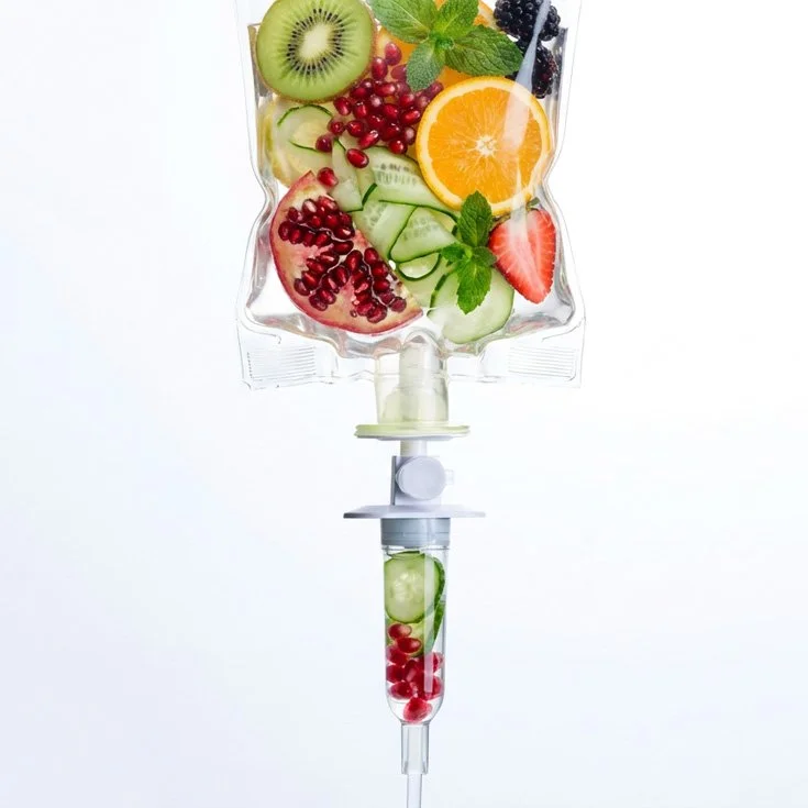 A medical IV bag filled with sliced fruits like kiwi, orange, strawberries, cucumber, pomegranate seeds, and mint leaves.