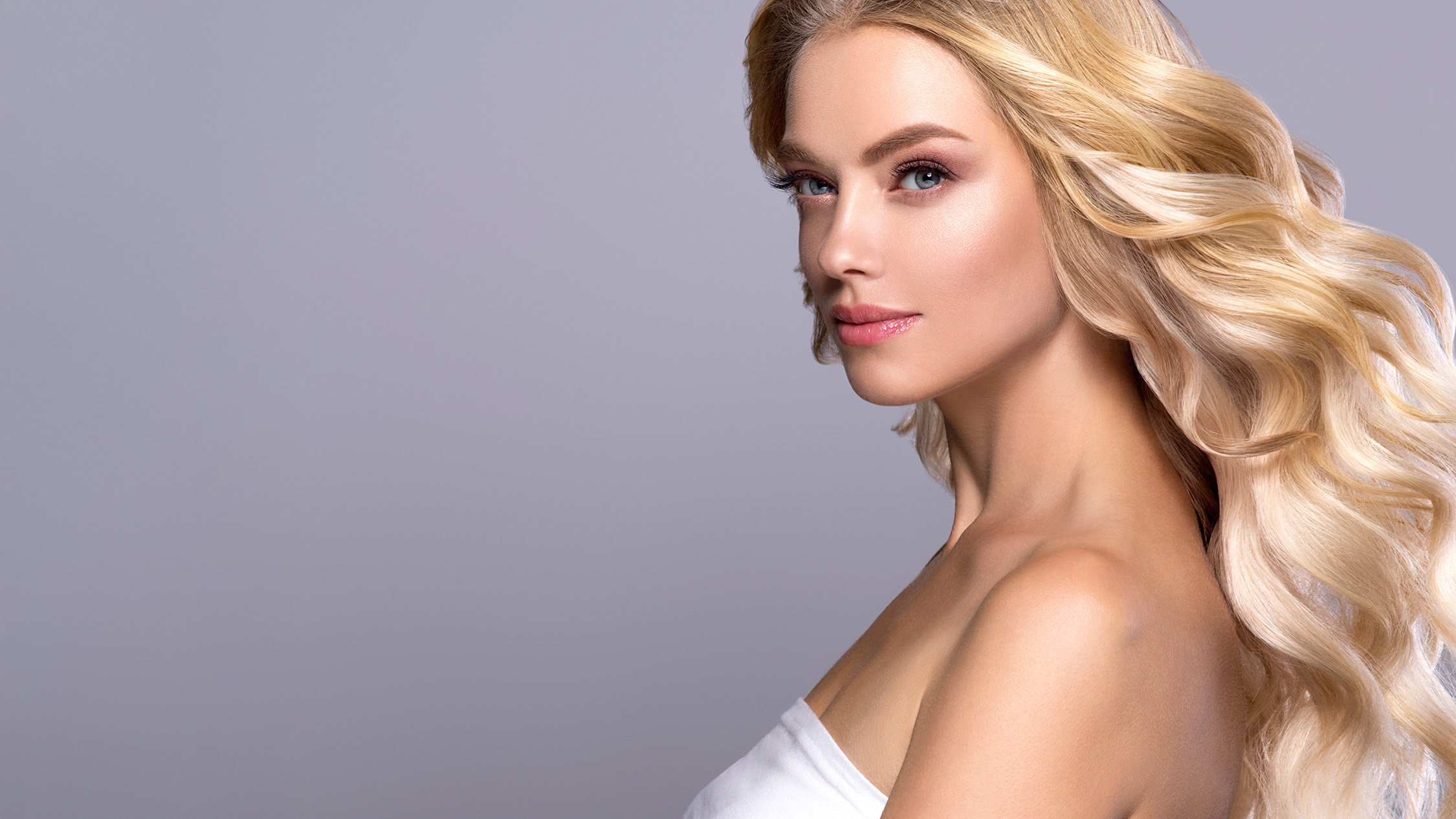 Close-up of a woman with long, wavy blonde hair, wearing a white top, against a plain gray background.