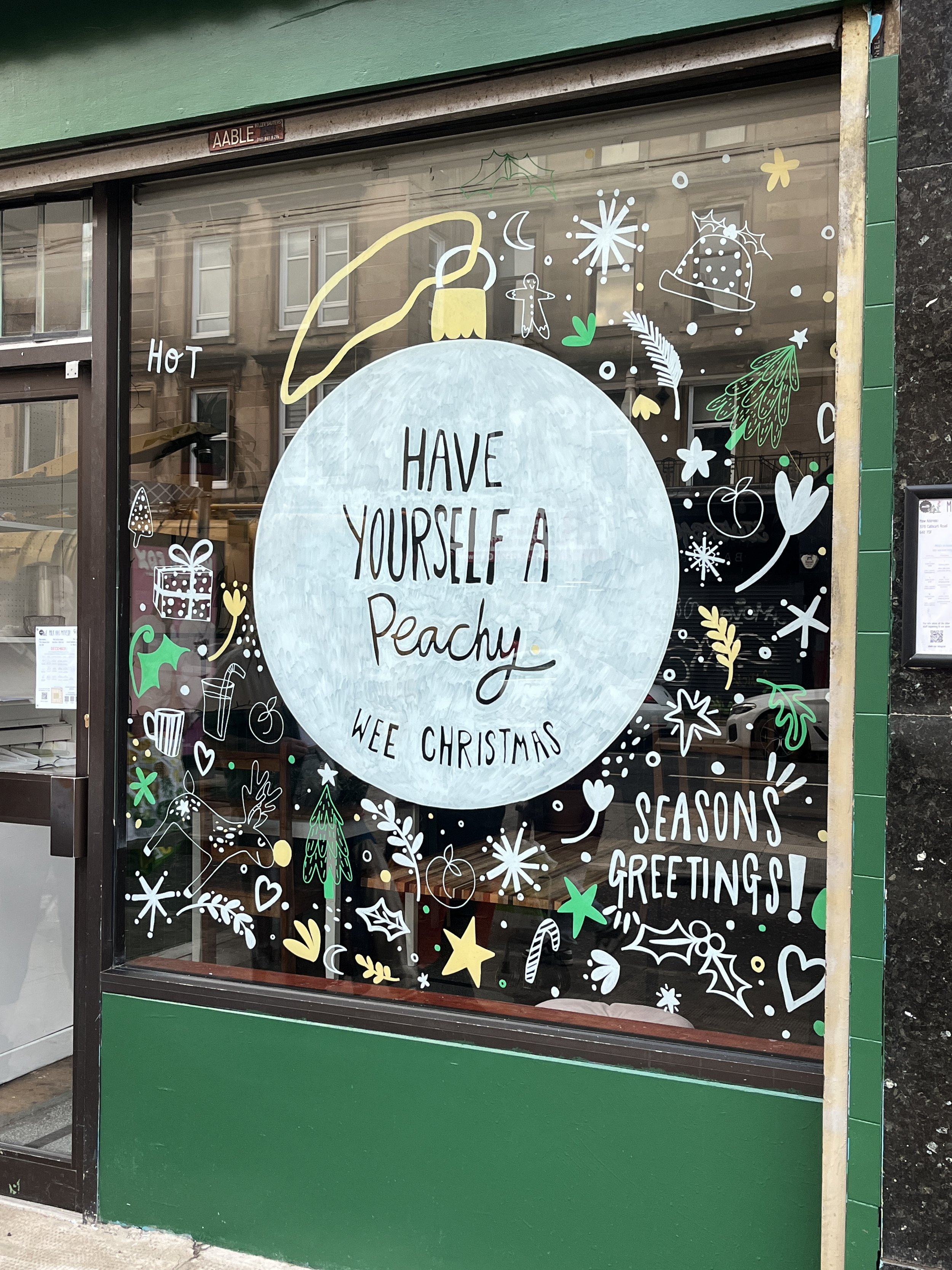 Storefront window decorated with holiday illustrations and a large ornament shape, with the message "Have yourself a peachy wee Christmas" and "Seasons greetings!" written on it. Decorative elements include trees, snowflakes, stars, holly, candy canes, hearts, a reindeer, gifts, and festive symbols in white, yellow, green, and blue.