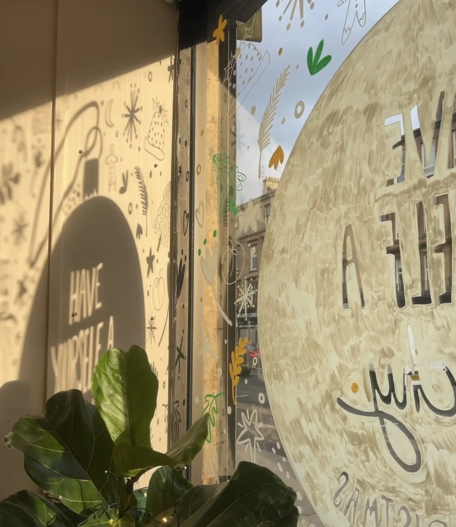 Decorative window with painted designs and plants outside, reflecting sunlight and showing part of a large circular sign with text.