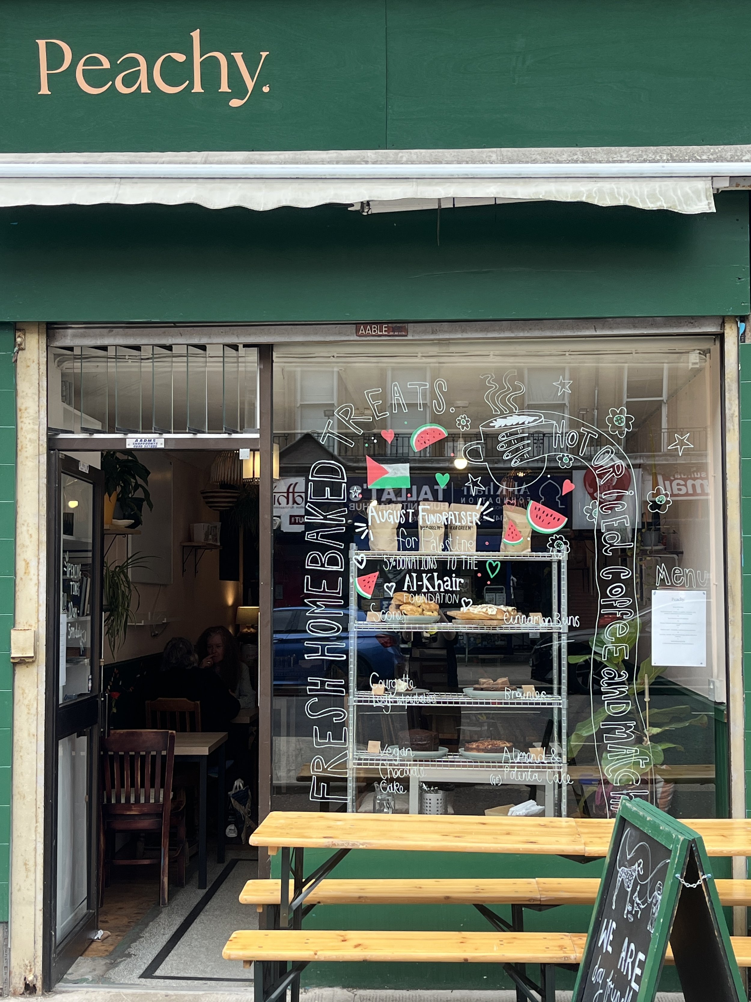 The storefront of a restaurant named Peachy with outdoor seating, a menu board, and a glass window displaying the menu items inside. The window also features colorful illustrations and text promoting baked treats and coffee.