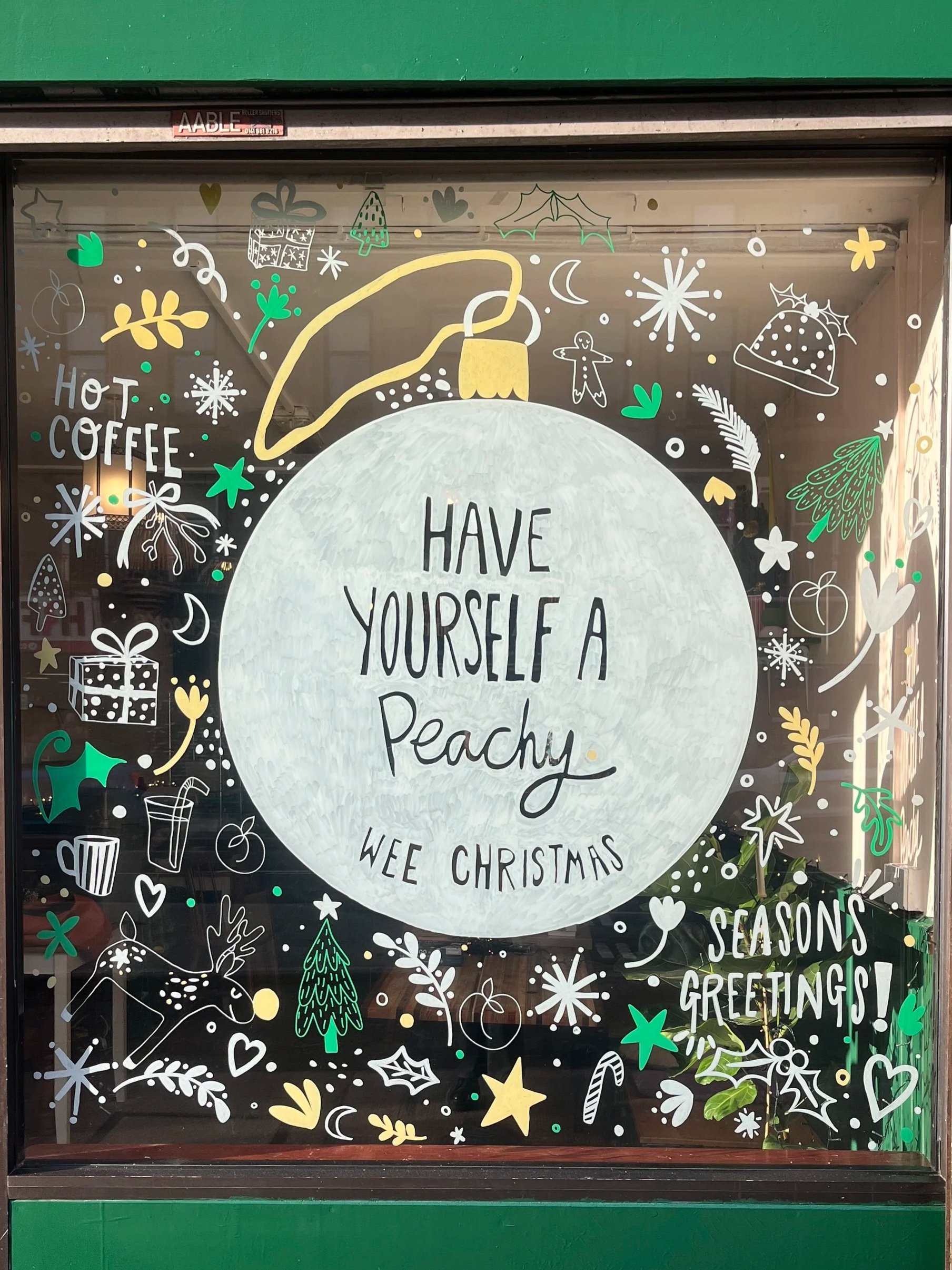Holiday window display with a large white ornament decorated with the message 'Have yourself a peachy wee Christmas.' Surrounding the ornament are various festive illustrations, including gifts, trees, holly, stars, snowflakes, a reindeer, a gingerbread man, a hat, and other holiday-themed doodles, with the words 'SEASONS GREETINGS!' on the right side of the window.