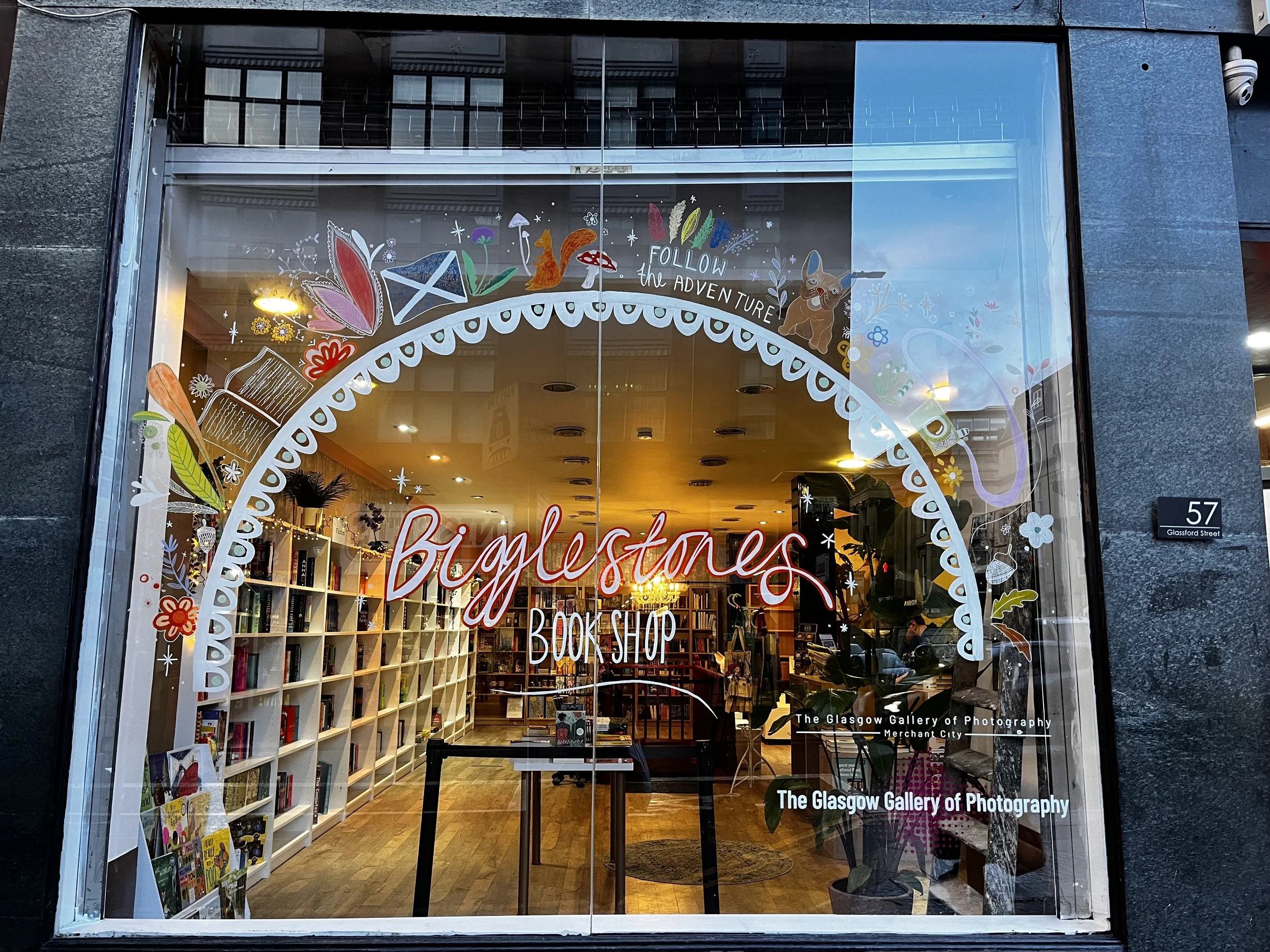 The front window of a bookstore called Biglessones Book Shop with colorful decorative illustrations and text on the glass, including flowers, books, and the phrase 'Follow the Adventure'. Inside, bookshelves and plants are visible.