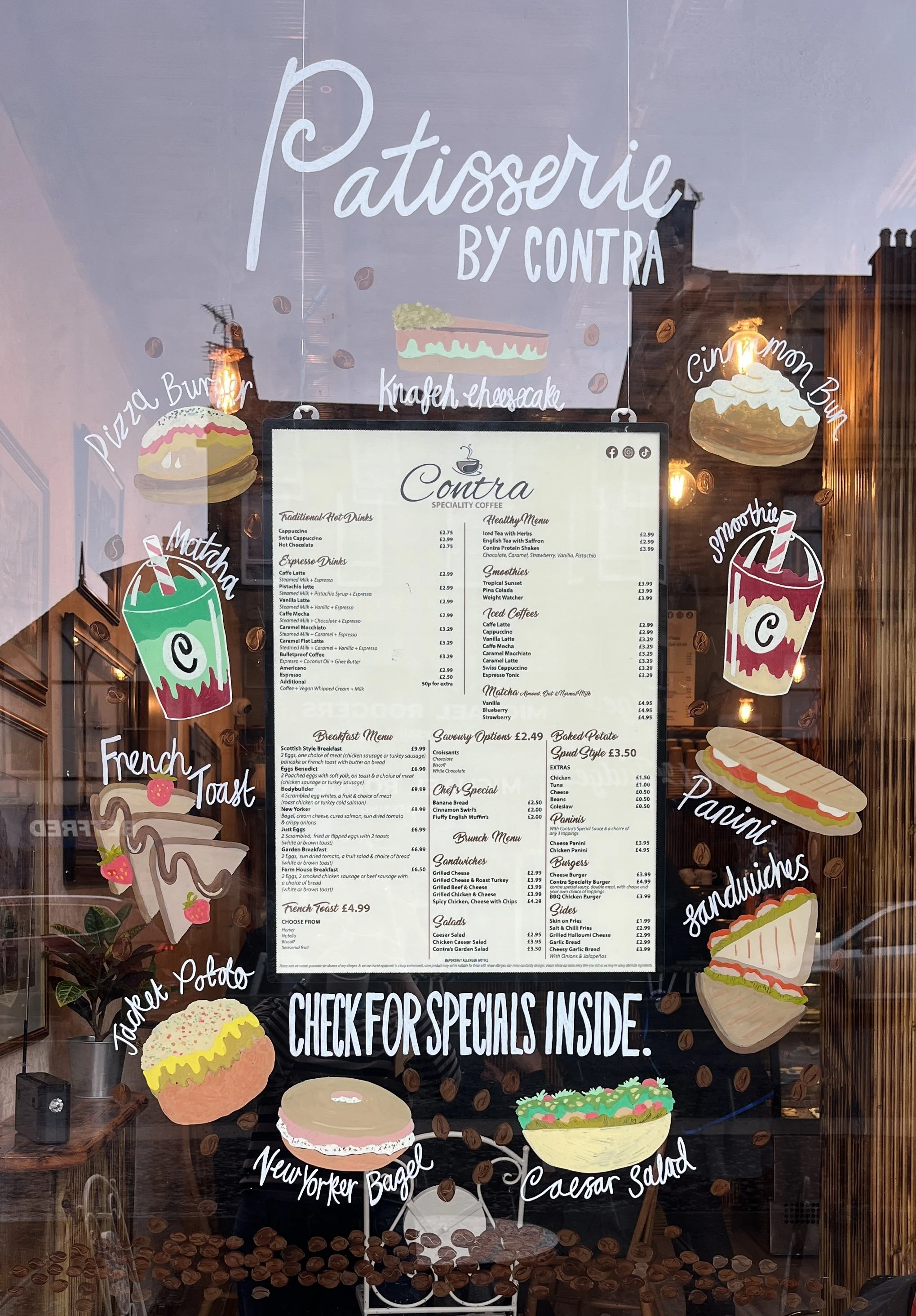 A window display of a bakery with colorful illustrations of desserts and food items, including a pizza bun, cinnamon bun, smoothie, French toast, jacket potato, New Yorker bagel, and Caesar salad, along with a menu listing various food and drink options.