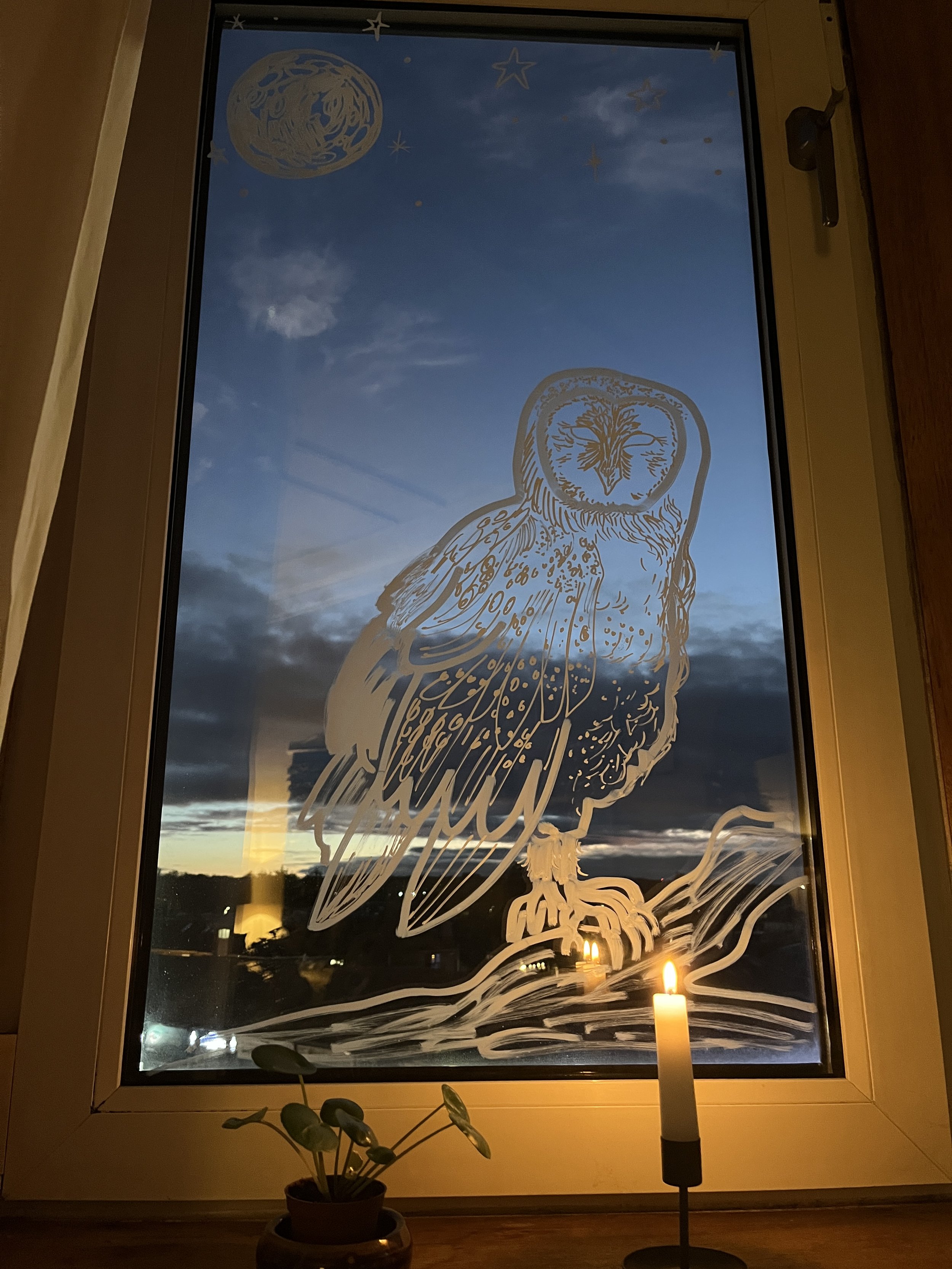 Decorative owl window sticker with a moon, stars, and clouds painted on a window, with a candle and plant below during dusk.