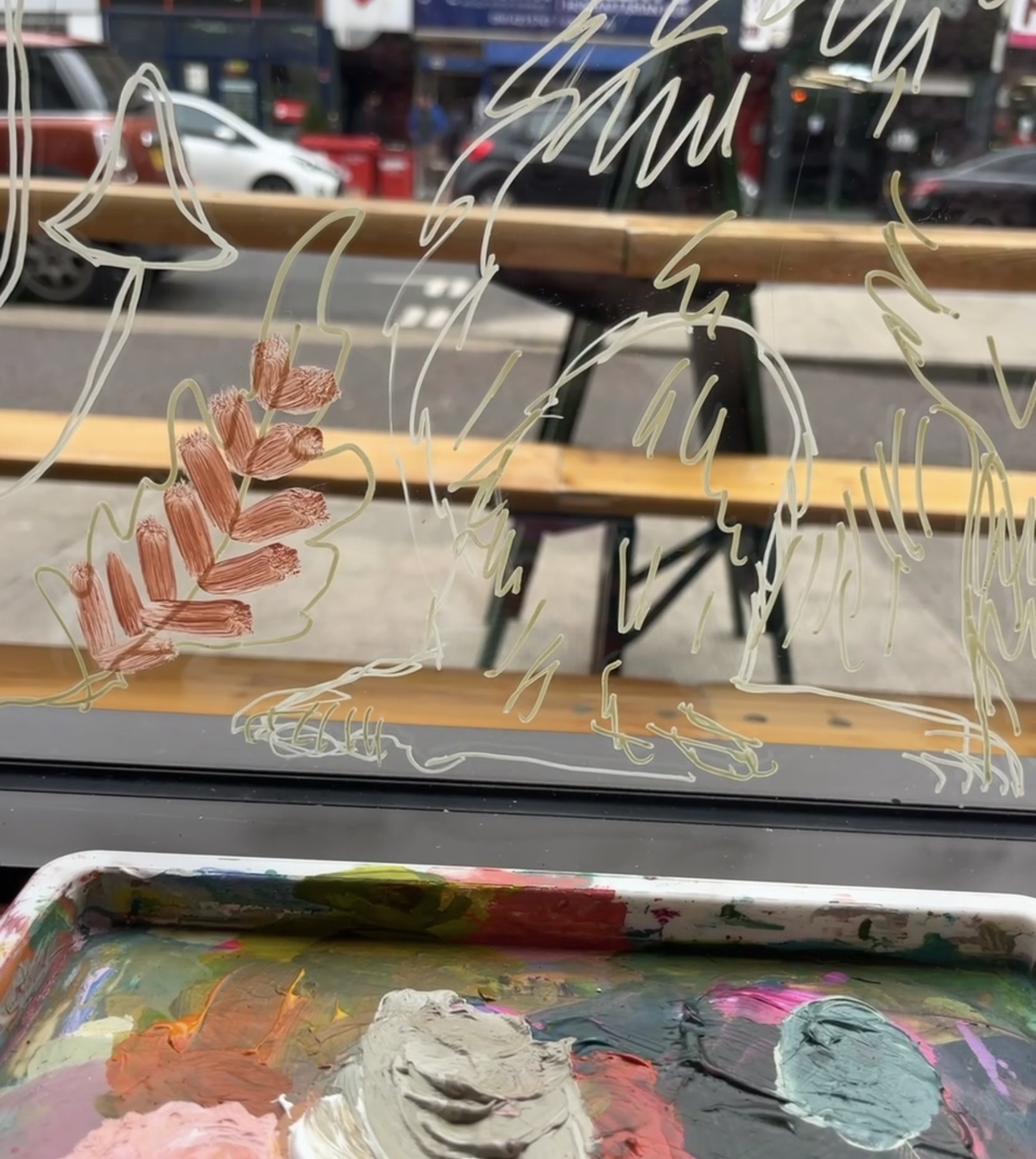 View through a window with painted and drawn designs of floral patterns in white and red, with a wooden table and a street scene visible outside, including parked cars and buildings.