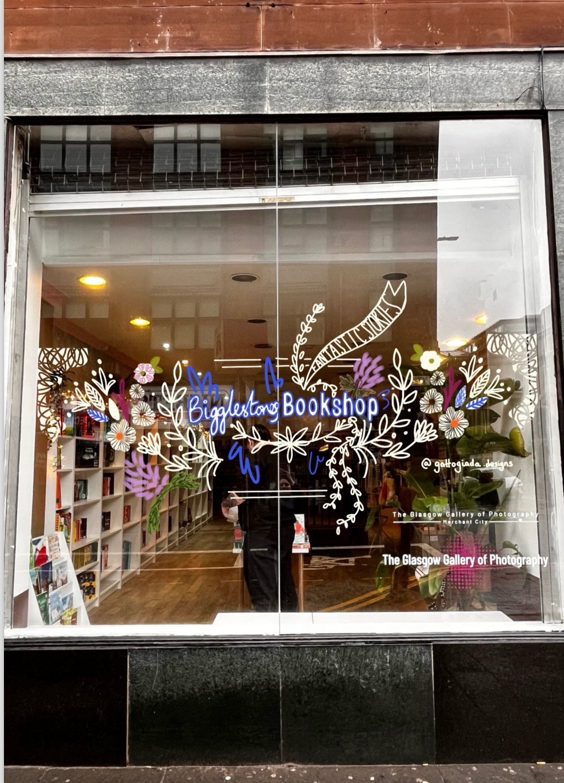 Storefront window of Big Glass Bookshop with decorative white floral and leaf design, blue neon sign, and various books visible inside.