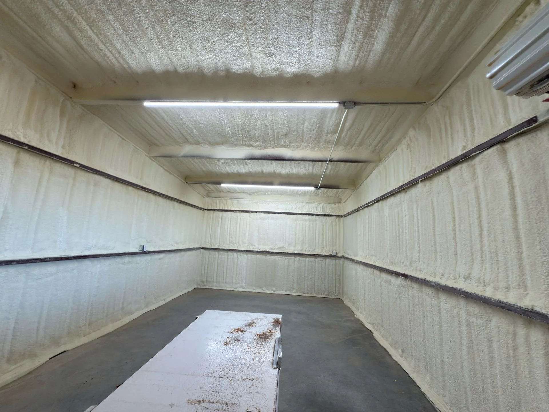 Empty indoor room with beige spray foam insulation on the walls and ceiling, exposed wiring, a metal table with rust spots, and fluorescent lighting.