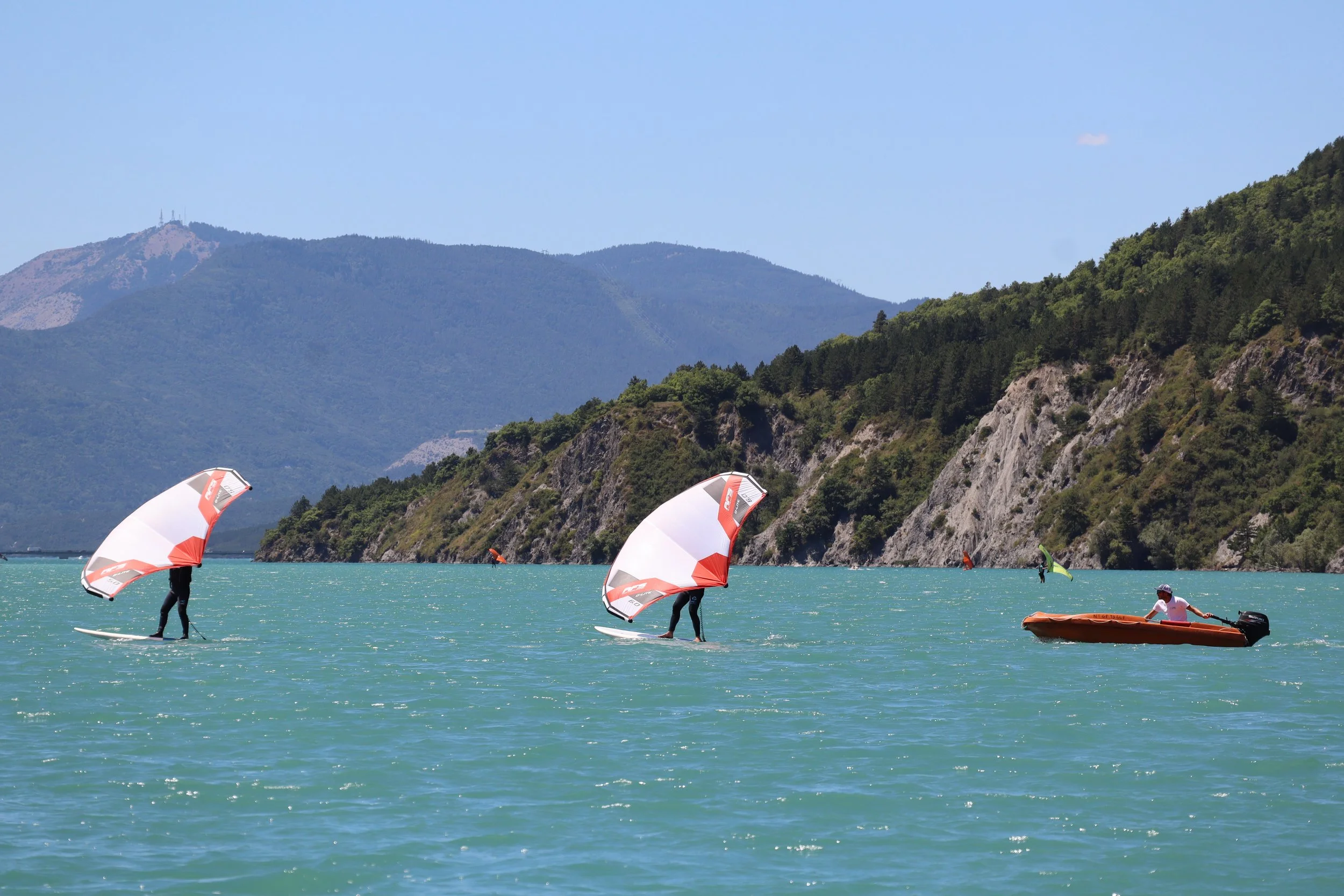 Trois personnes pratiquent la planche à voile sur un lac avec des montagnes en arrière-plan, et une personne en bateau à moteur.