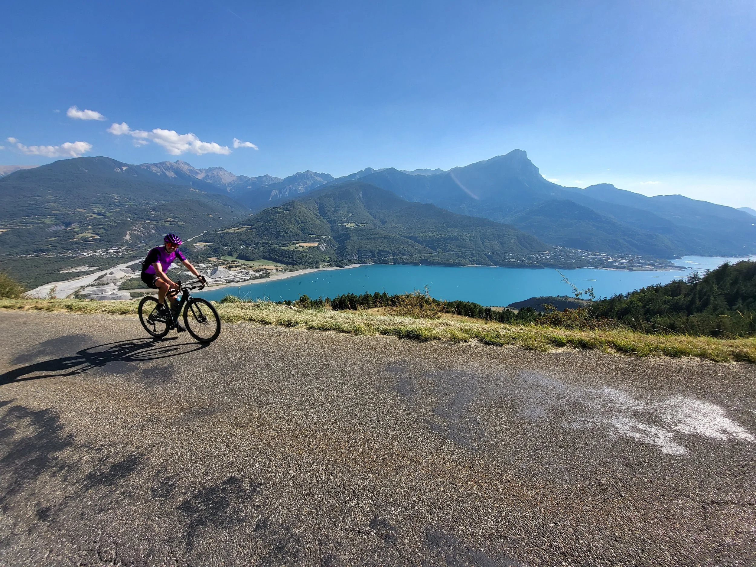 Cycliste en vélo noir et violet sur une route de montagne avec vue sur un lac bleu, entouré de montagnes et ciel bleu