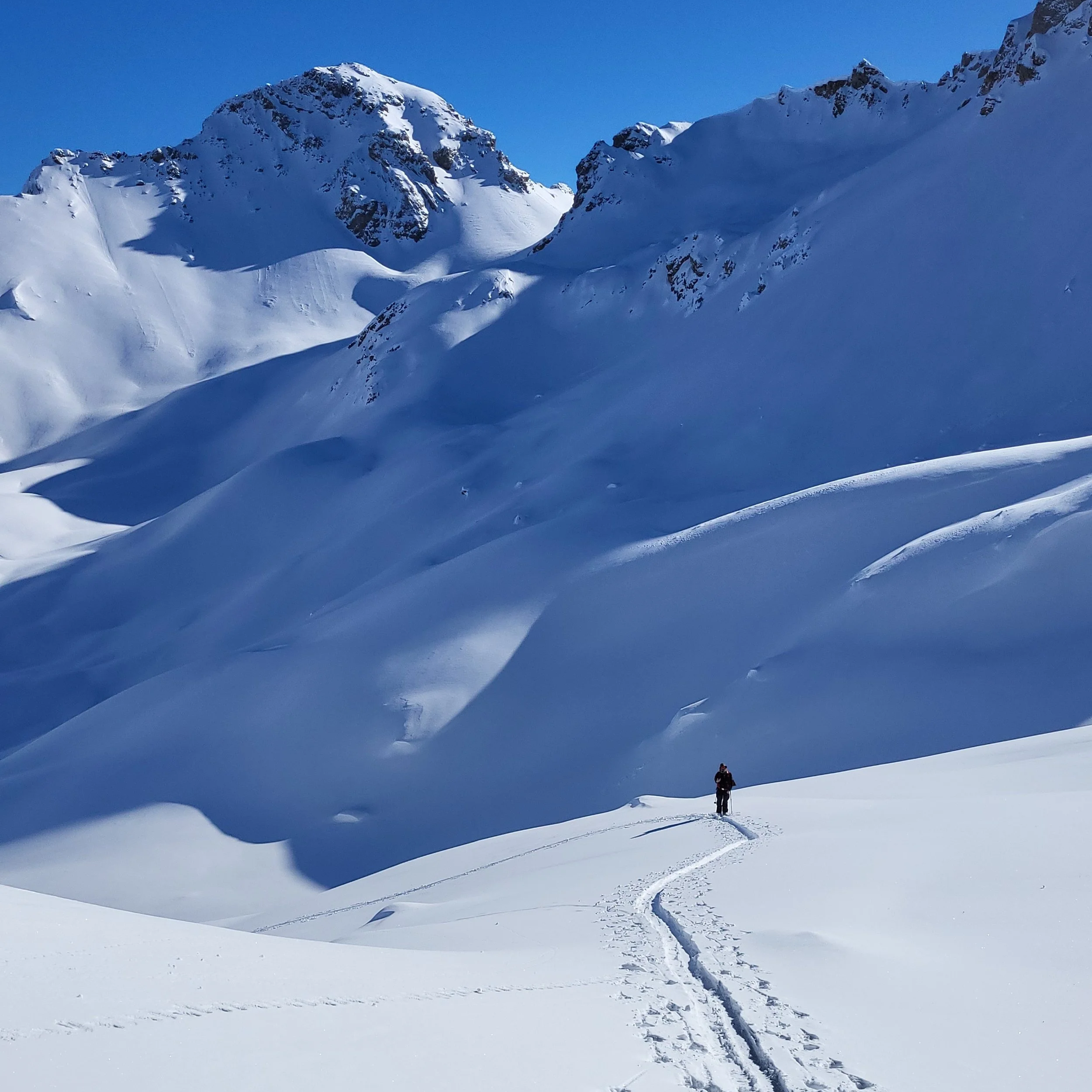 Une montagne enneigée avec un ciel clair. Un seul skieur ou randonneur marche dans la neige, laissant une trace derrière lui.