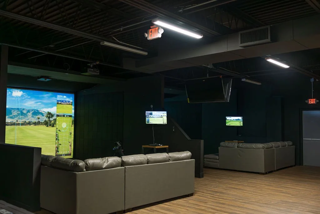 Indoor golf simulation room with large screen displaying a golf course, two wall-mounted TVs, leather sofas, a small table, and an emergency exit sign.
