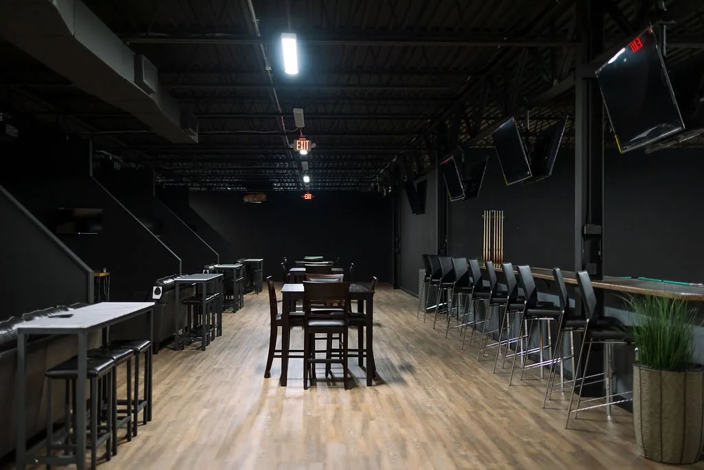 Empty bar or lounge area with wooden flooring, dark walls, bar stools along a counter, high tables with chairs, multiple ceiling-mounted TVs, and plants for decor.