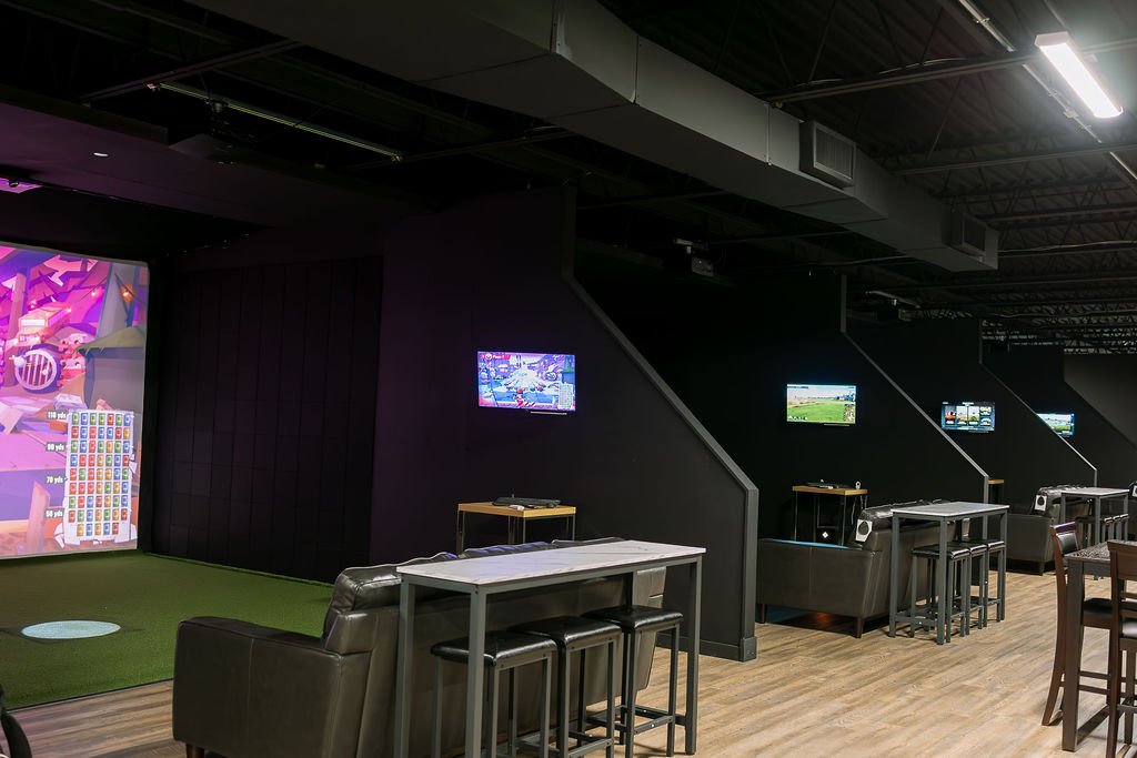 A gaming lounge with multiple gaming stations, each equipped with a TV, speaker, and seating area. The space has a modern design with black walls, wooden floors, and dim lighting.