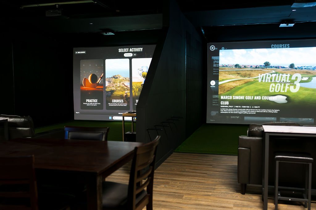Indoor golf simulator room with two large screens displaying virtual golf courses and activities, and seating area with tables and chairs.