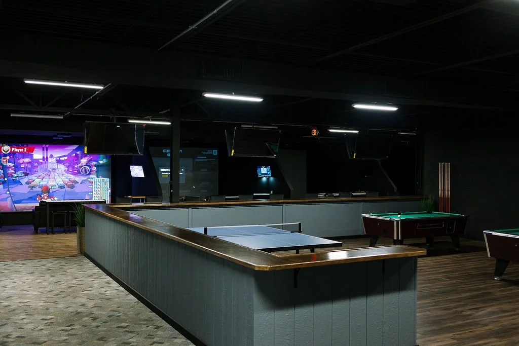 An indoor entertainment area featuring a pool table, air hockey table, and arcade gaming consoles with large screens, in a dimly lit room.