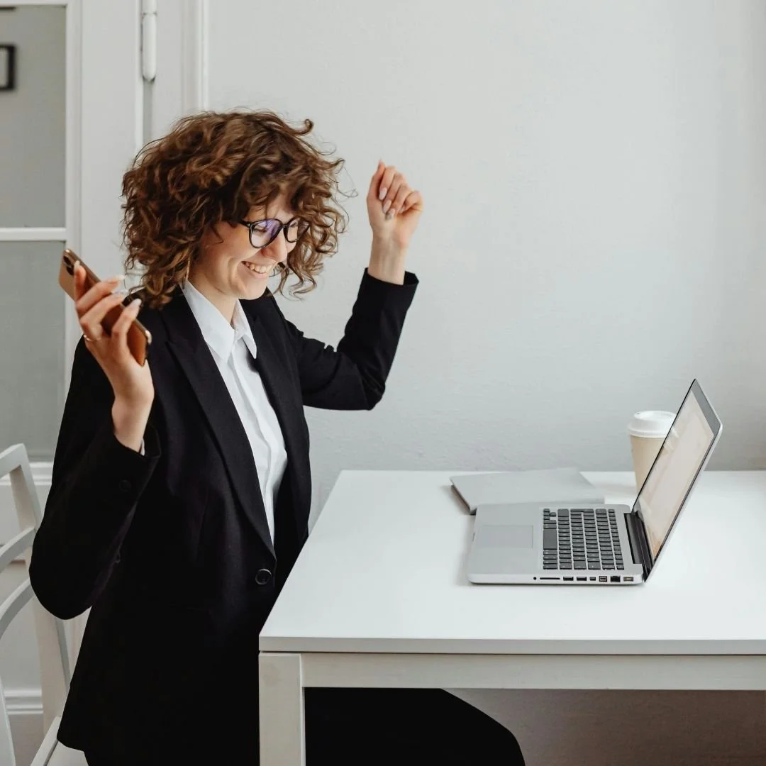 A woman in a black blazer and white shirt, with curly hair and glasses, happily dancing in front of a laptop on a white desk in an office.