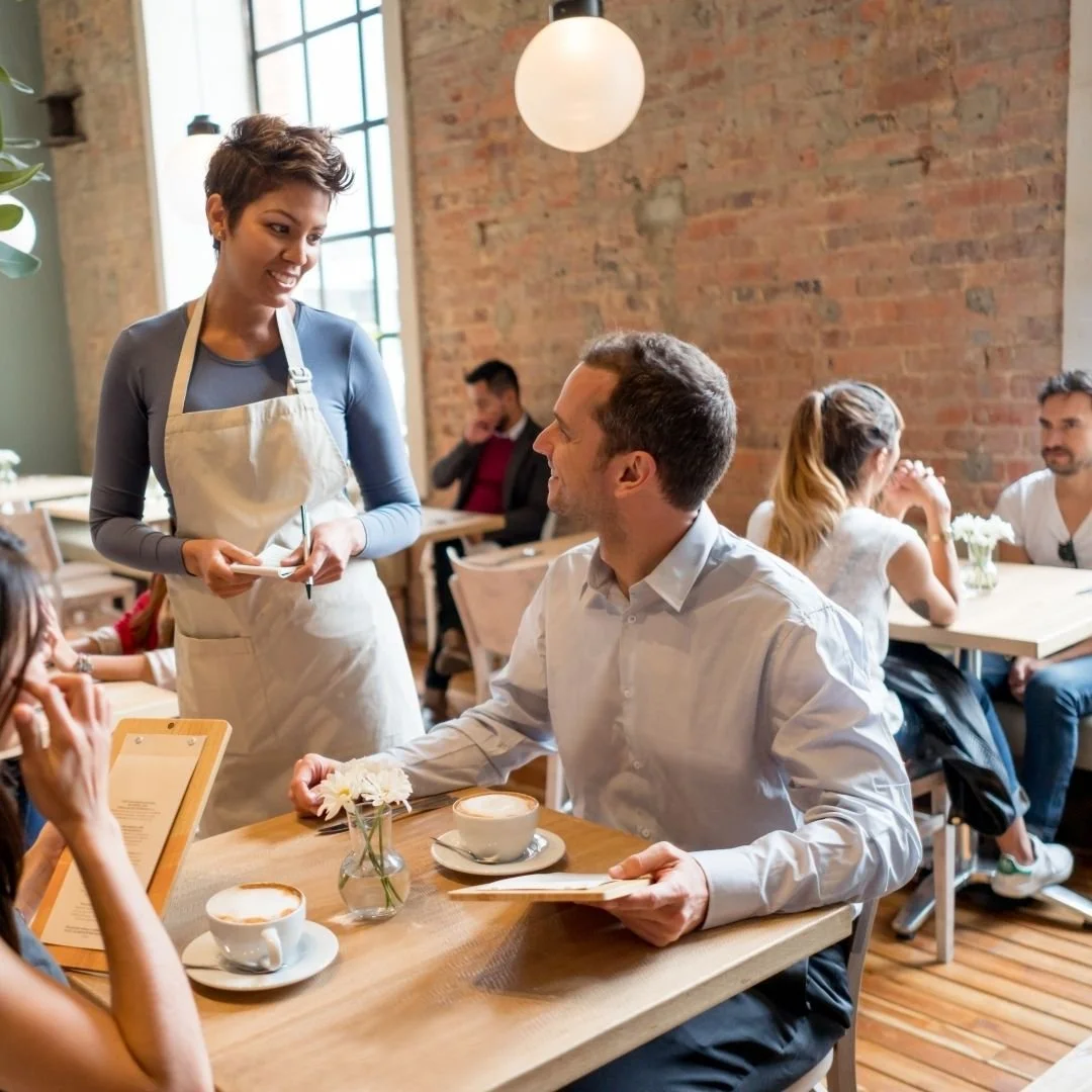 A waitress taking an order from a man sitting at a table in a cozy cafe with a brick wall and large window.