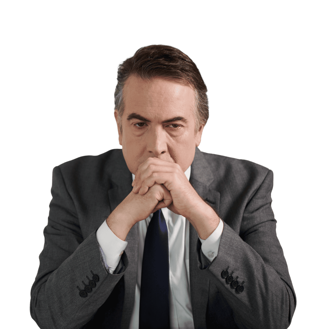 A middle-aged man in a dark gray suit with a white shirt and black tie, sitting with his hands clasped near his face, looking directly at the camera with a serious expression.