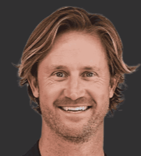Close-up of a smiling man with medium-length brown hair and a beard, wearing a dark shirt.