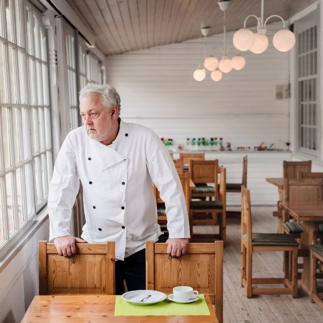 A sad chef in a white uniform standing alone at a table in a bright, empty restaurant, with napkin, plate, and cup on the table.