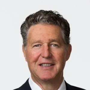 Close-up portrait of a middle-aged man with curly dark hair, wearing a suit and tie, smiling against a light gray background.