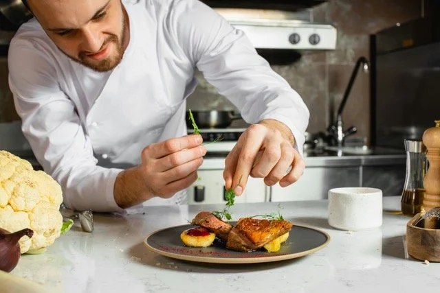 Chef garnishing a plated dish of grilled meat with herbs in a modern kitchen.