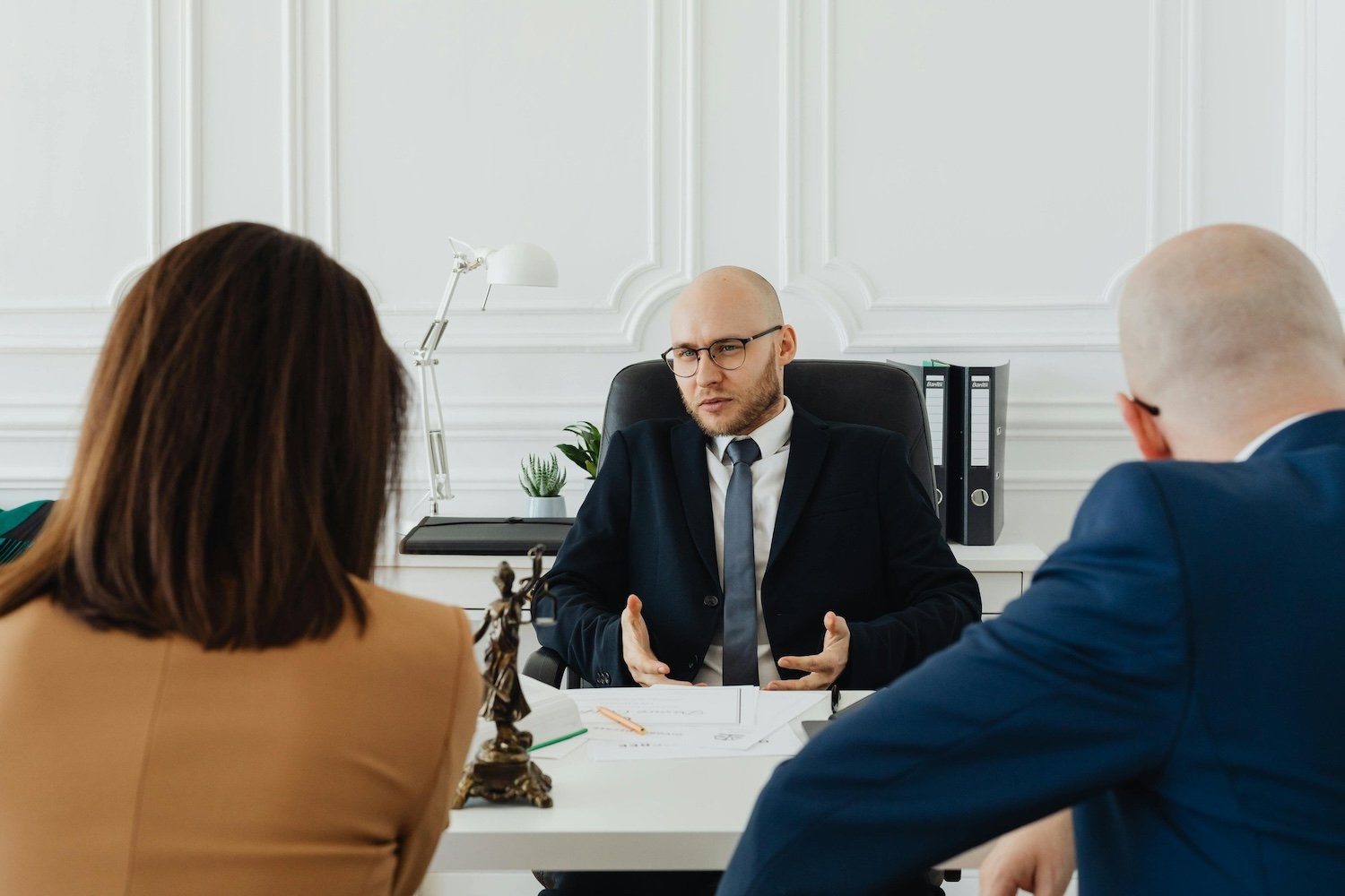 A man in a suit and glasses is talking in a meeting with two people, a woman and a man, in an office with white walls and a desk.