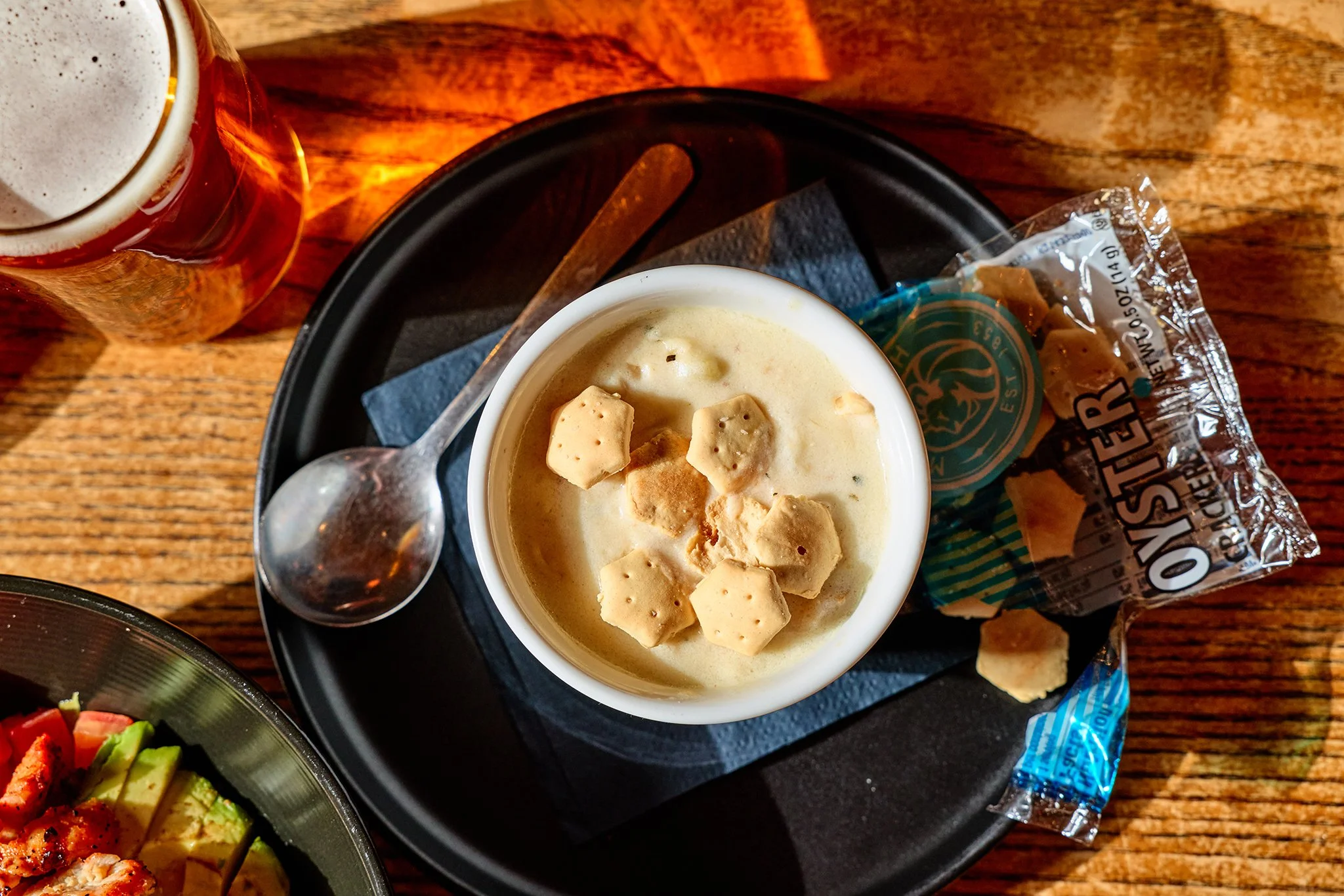A bowl of creamy soup topped with oyster crackers, placed on a black tray with a spoon, next to a bag of oyster crackers and a glass of beer on a wooden table.