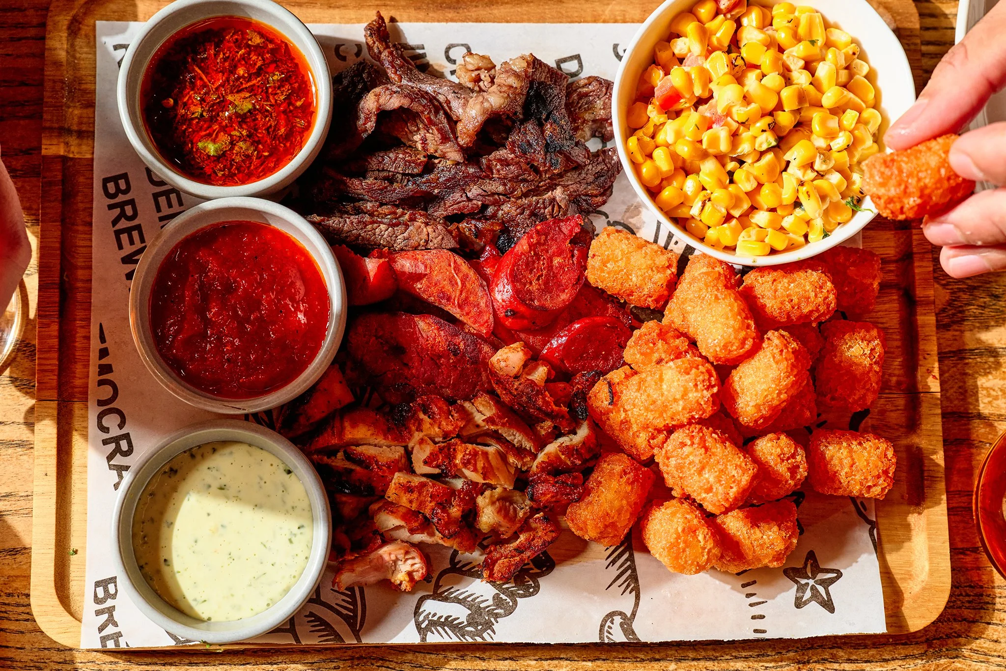 A tray of mixed fried foods including breaded mozzarella sticks, grilled meats, potato chips, and assorted sauces with a bowl of corn and a bowl of salsa.
