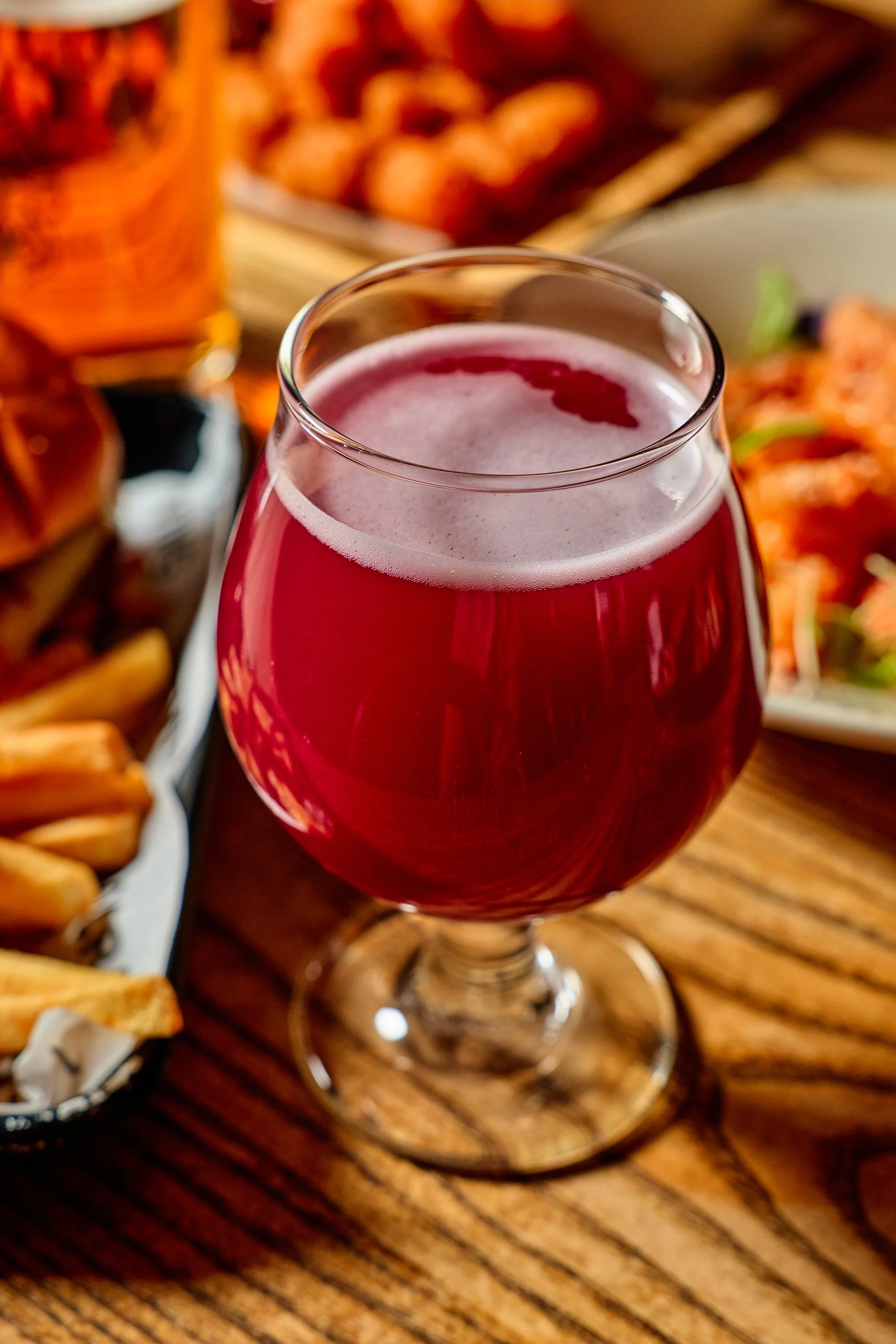 A glass of red craft beer with a foamy head on a wooden table, surrounded by fried foods and appetizers.