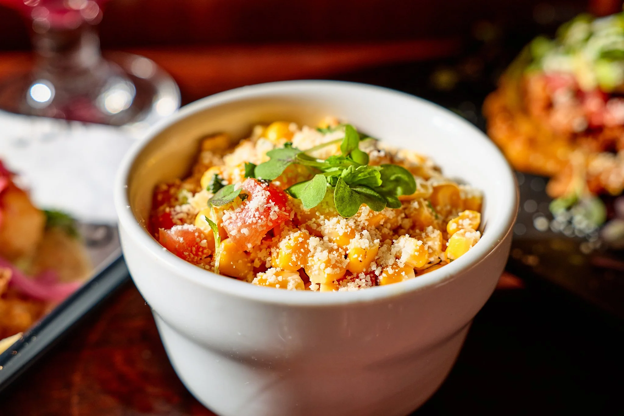 A white bowl filled with a corn salad topped with shredded cheese, tomato chunks, and fresh herbs.