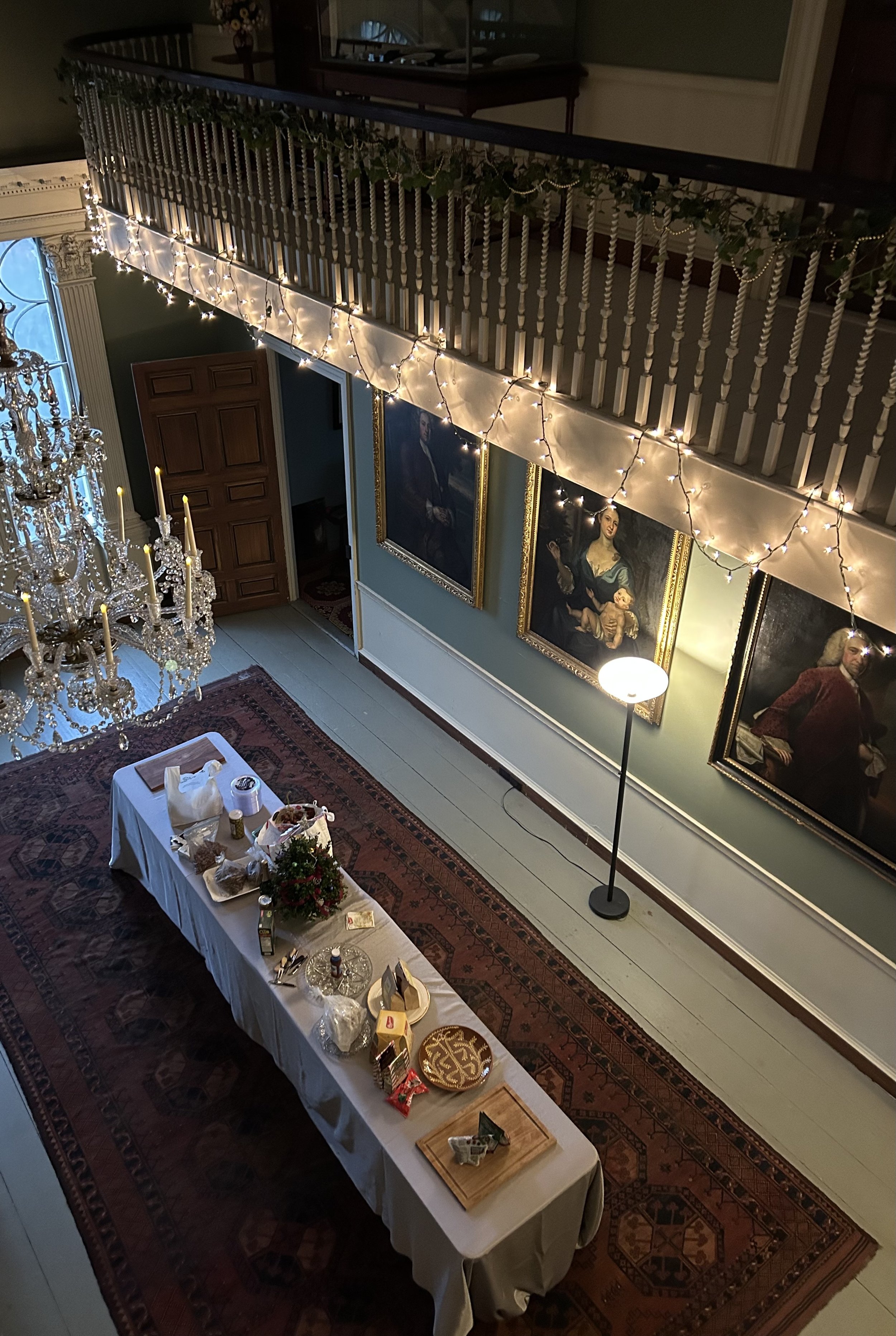 Interior of a house decorated for a celebration with a long table set with food and decor, a chandelier, framed portraits on the wall, a standing lamp, string lights, a staircase with a garland, and a large carpet.